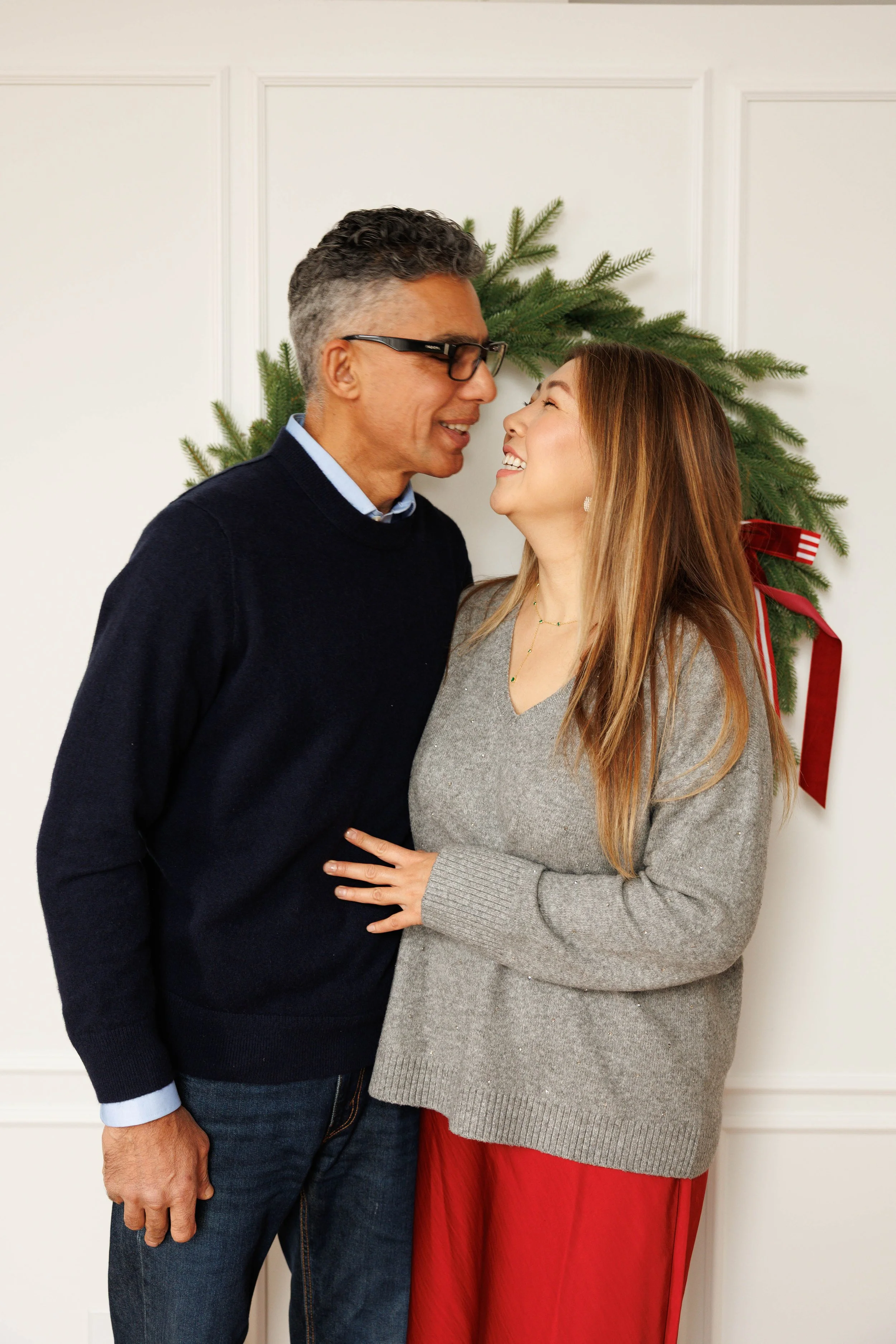 A happy couple smiling and looking at each other in front of a holiday wreath with a red bow on a white wall.