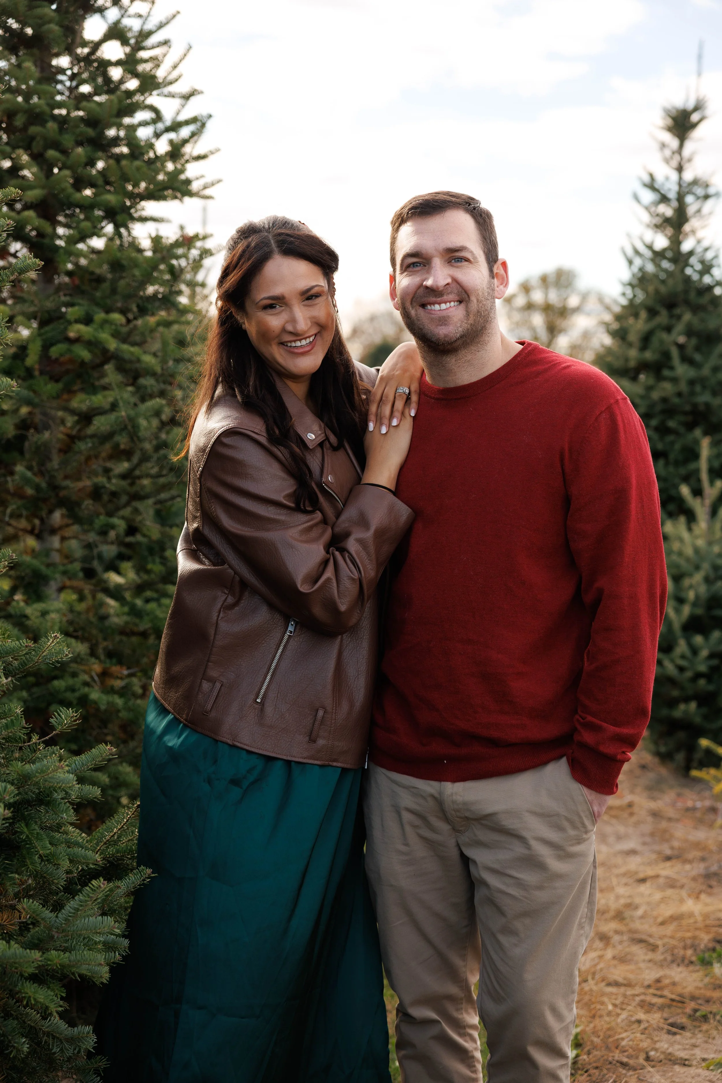 A smiling woman in a brown leather jacket and teal skirt, and a smiling man in a red sweater and beige pants, standing outdoors among evergreen trees.