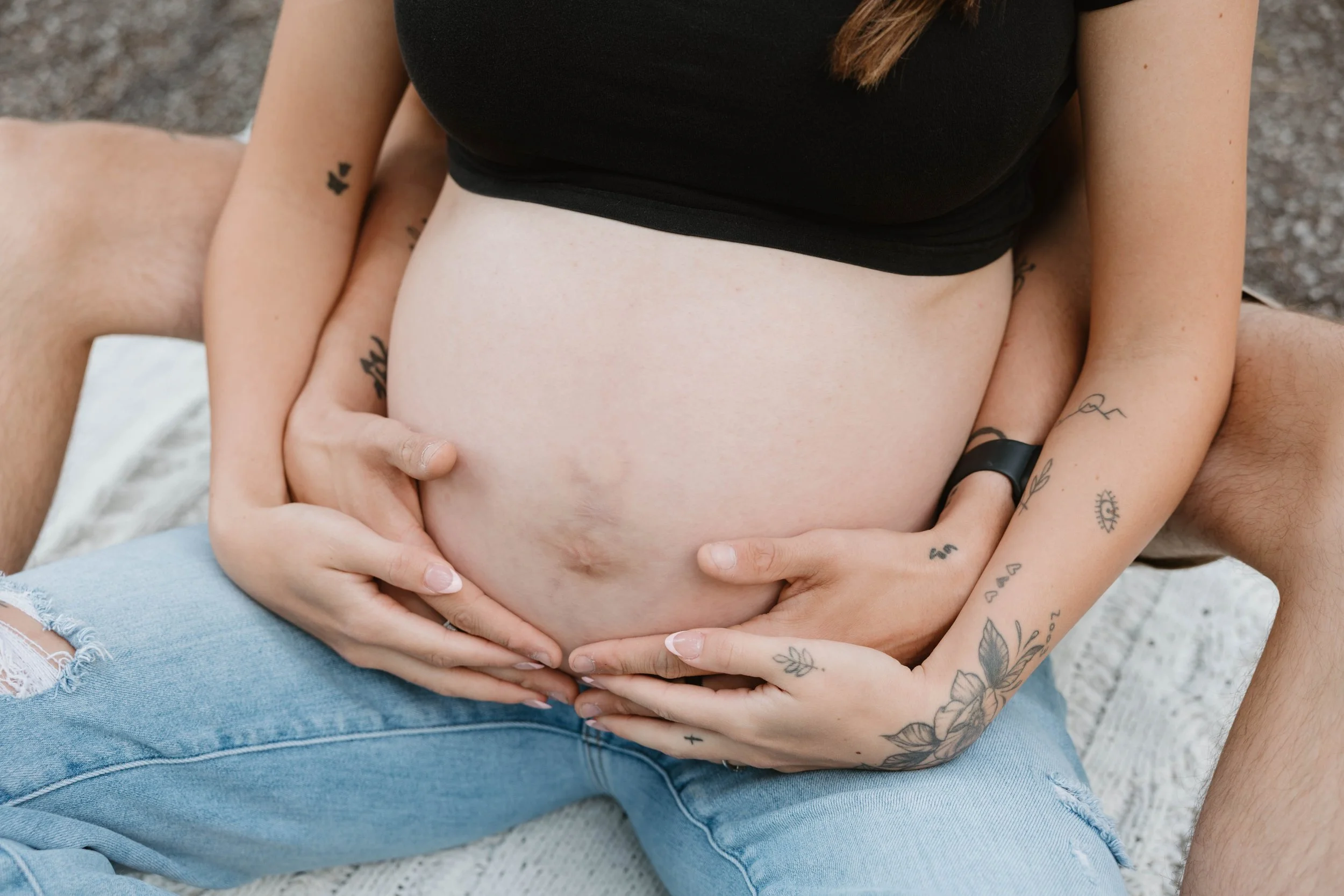 A pregnant woman with tattoos on her arms, wearing a black top and blue jeans, holding her belly while sitting on a blanket outdoors.
