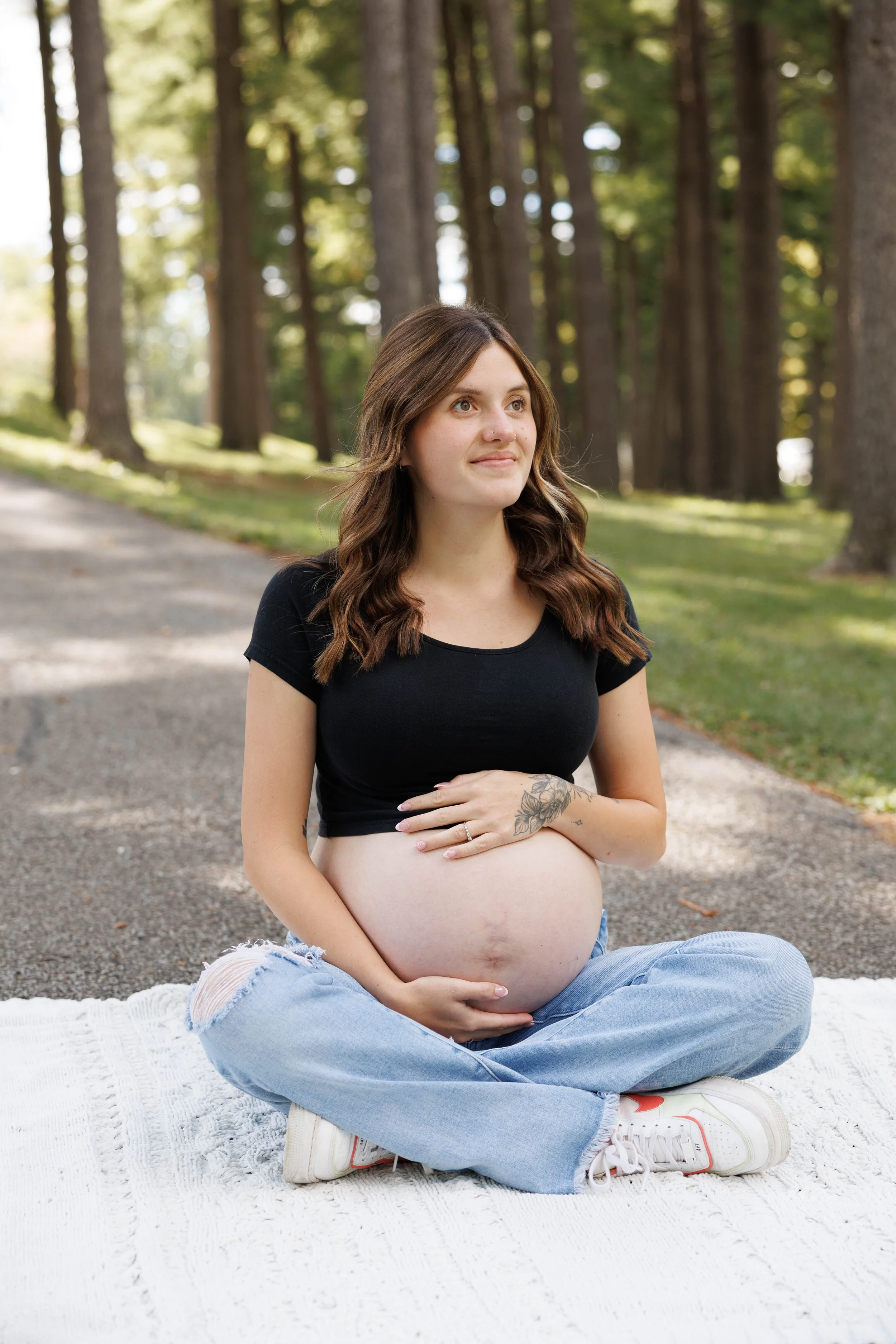 A pregnant woman with brown wavy hair sitting cross-legged on a white blanket outdoors in a wooded area, wearing a black crop top and ripped jeans, holding her baby bump.