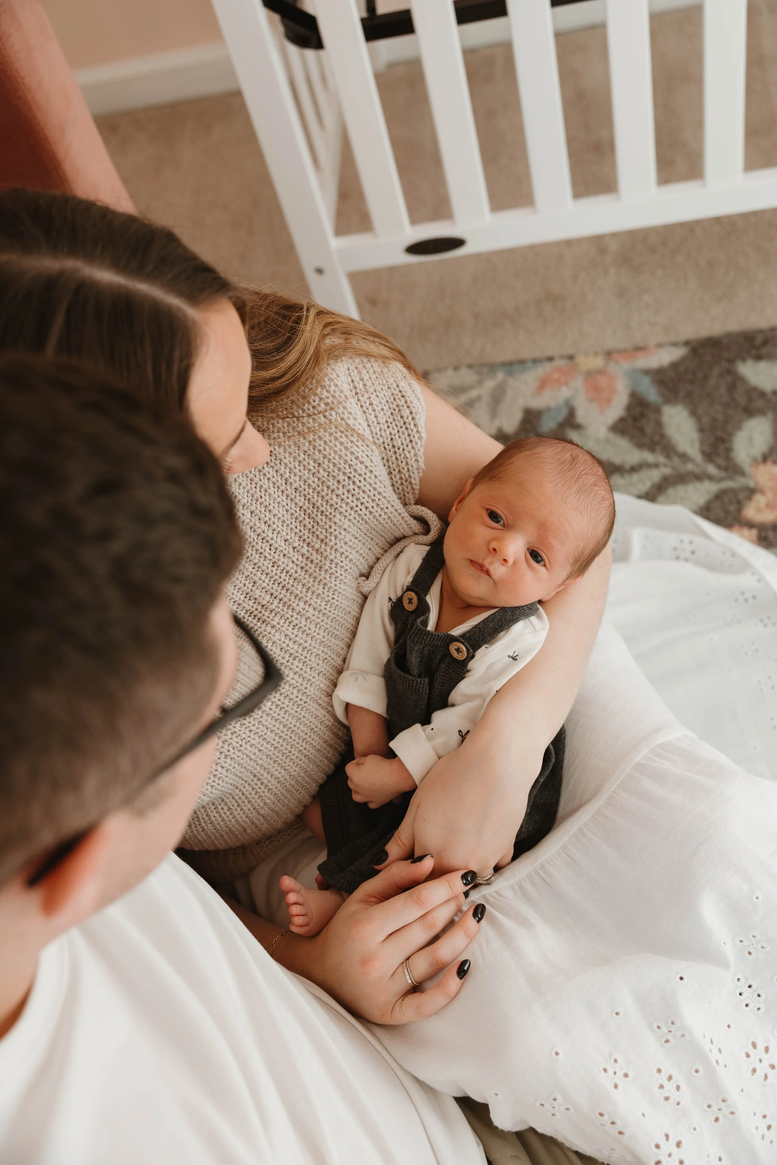 A newborn baby lying on a woman’s lap, being held and looked at by two adults, one wearing glasses, in a cozy room.