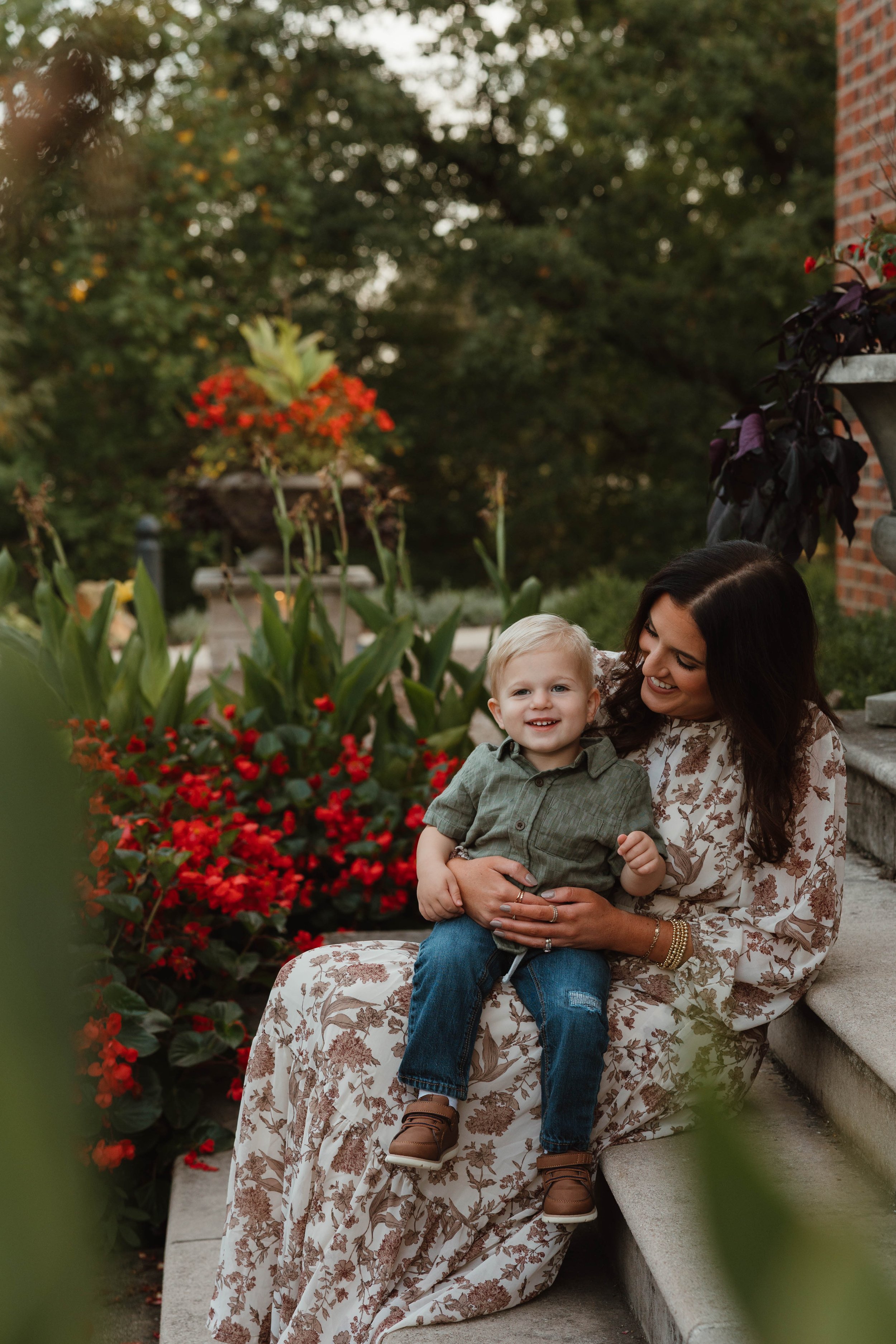 A woman sitting on outdoor steps holding a young boy, both smiling, with colorful flowers and greenery in the background.