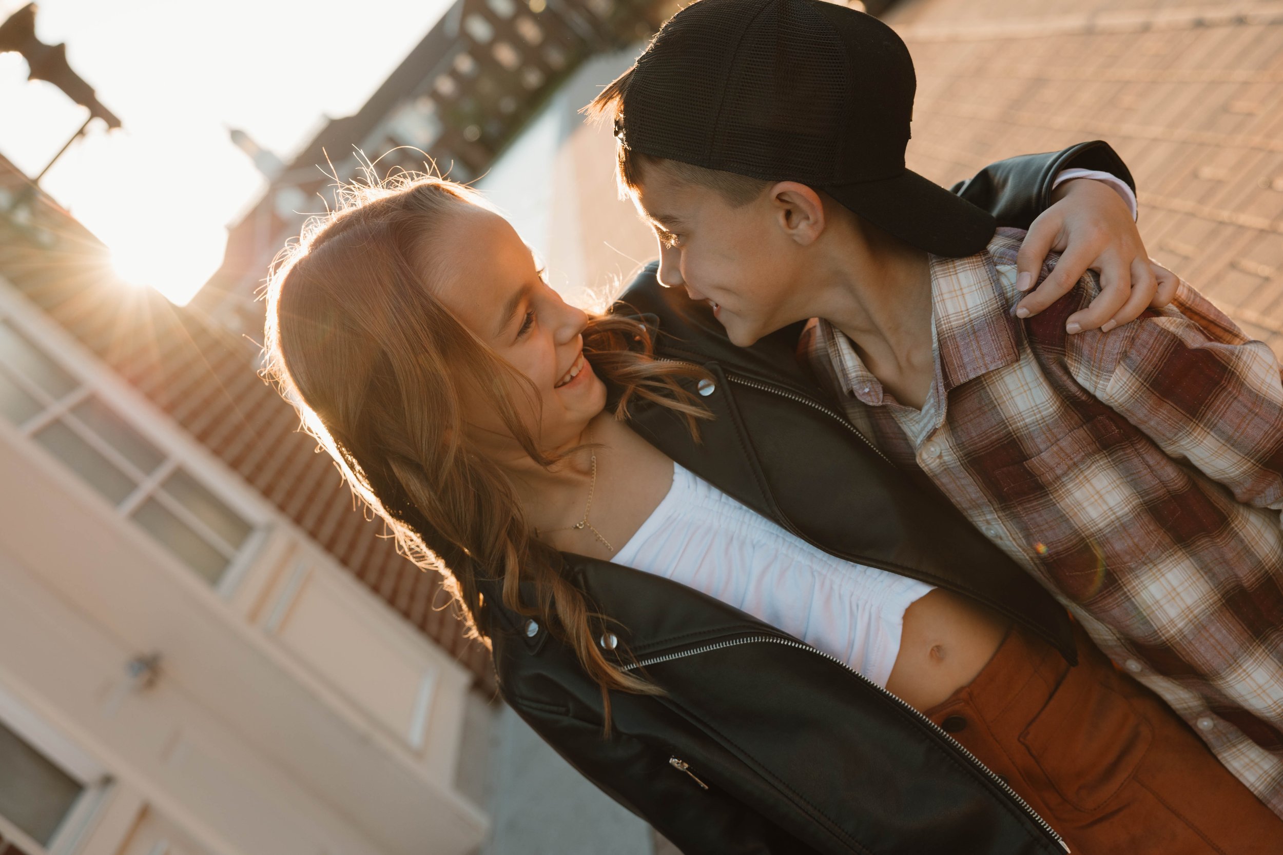 Young couple smiling and looking at each other while close together outdoors during sunset, with a brick building in the background.