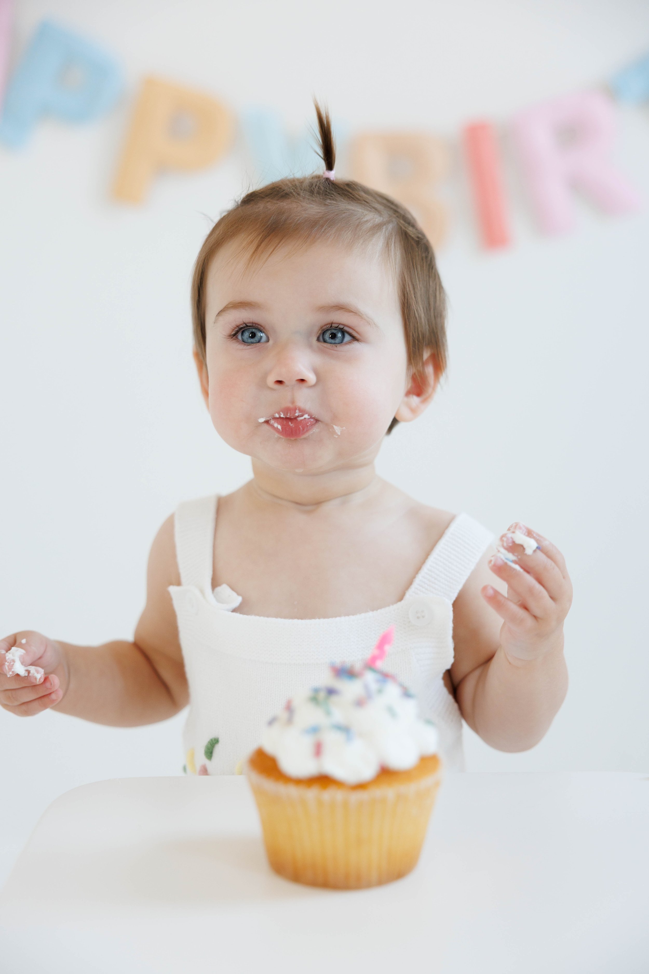 A young child with blue eyes and light brown hair tied in a small top knot, celebrating a birthday with a cupcake topped with whipped cream and a pink candle. The child has frosting on their lips and is holding a piece of cake in each hand.