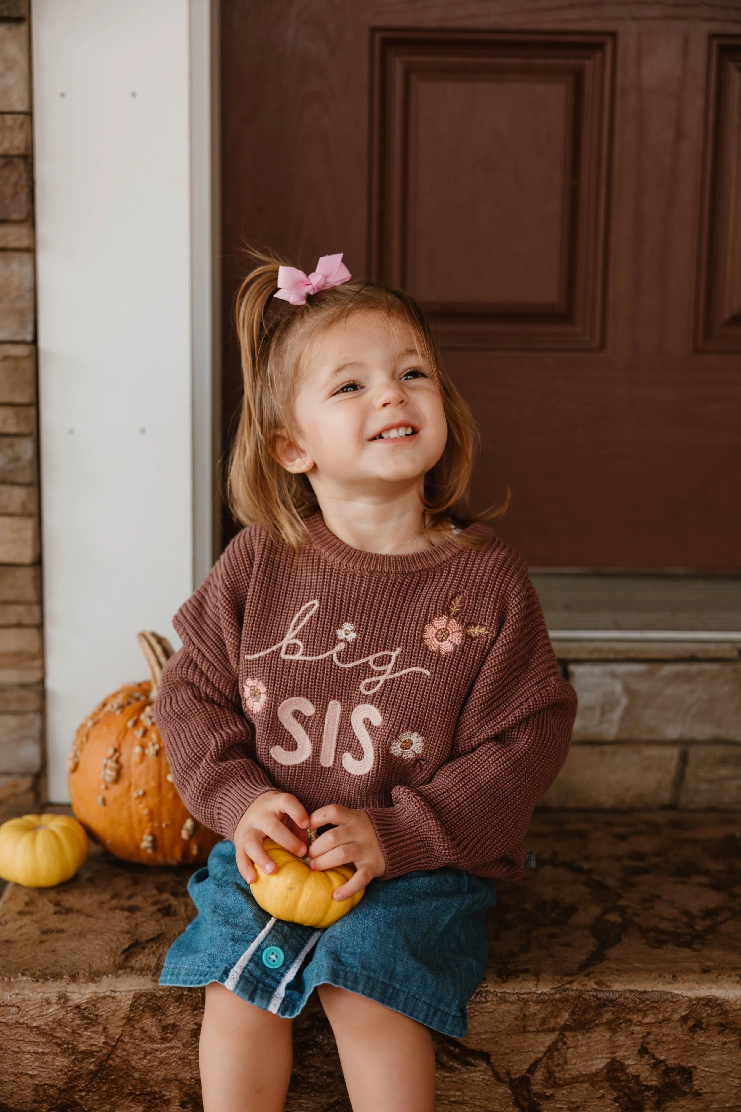 A young girl with a pink bow in her hair, wearing a brown sweater with embroidered flowers and words, sits on a stone step holding a small yellow pumpkin, with several larger pumpkins behind her on a porch.