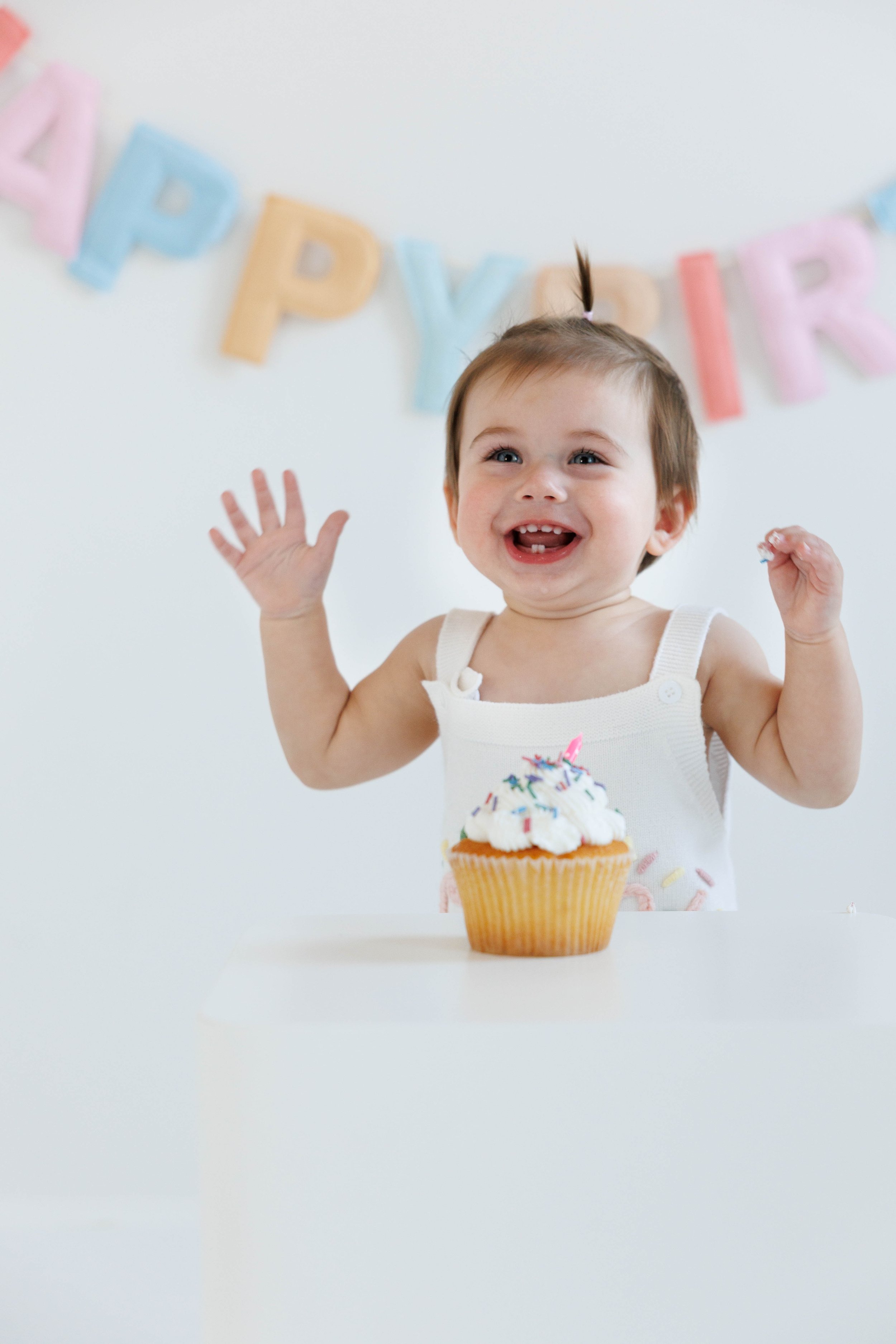 A smiling young girl at a birthday celebration, sitting in front of a cupcake with white frosting and colorful sprinkles, with a birthday banner in pastel colors in the background.