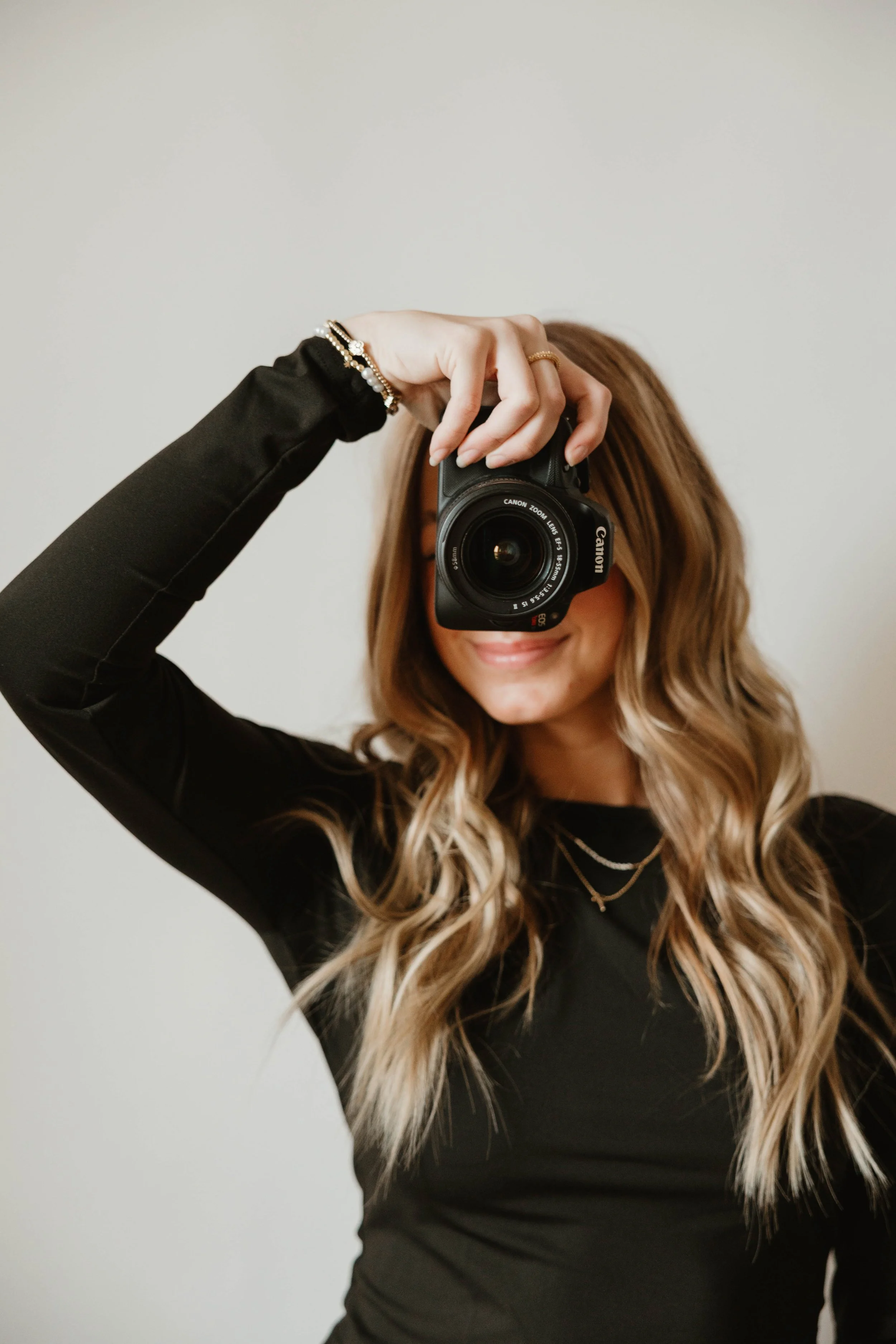Woman with blonde wavy hair taking a self-portrait with a Canon camera, smiling, wearing a black long-sleeve top, jewelry, and a necklace, standing against a plain background.
