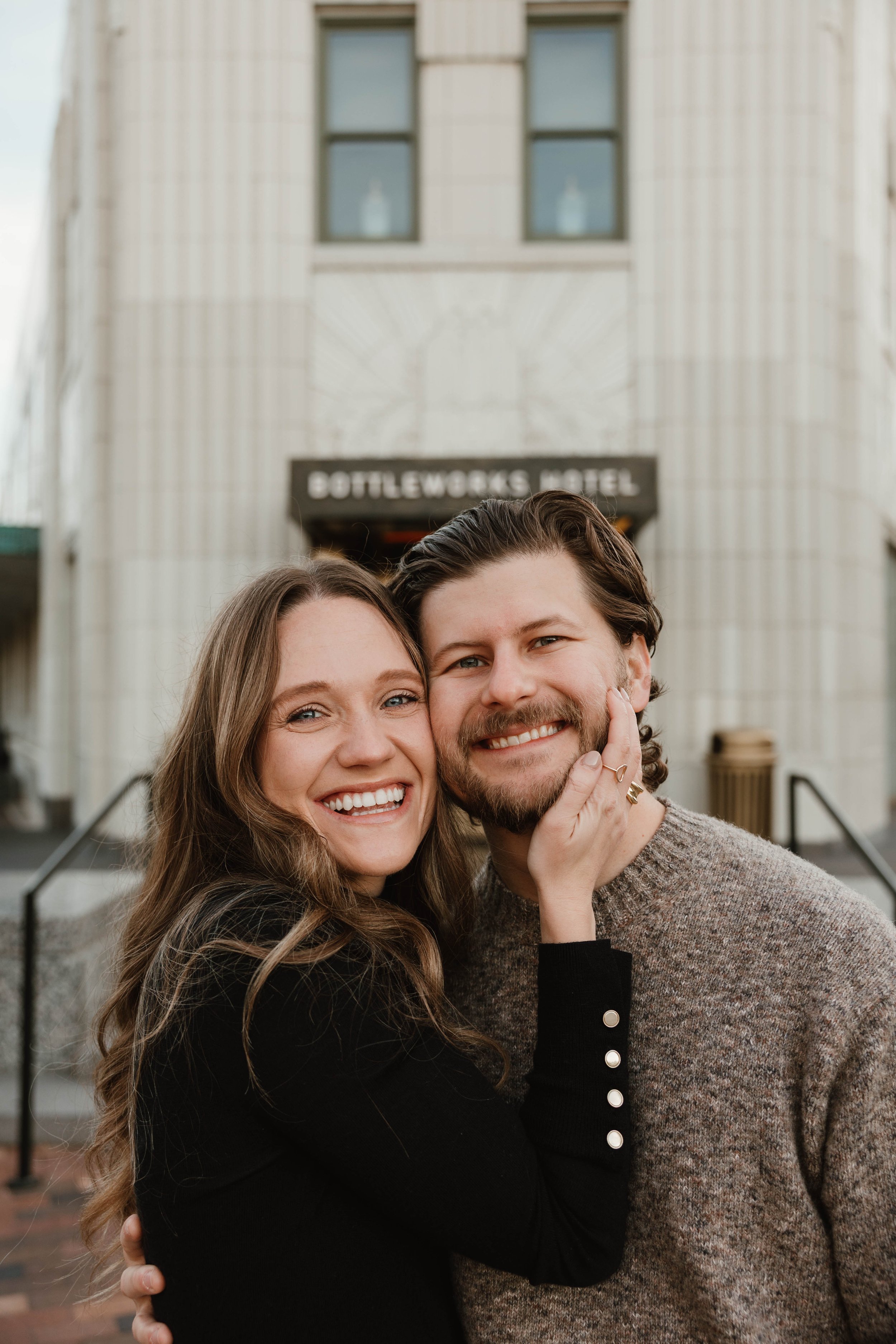 A smiling couple hugging in front of the Bottleworks Hotel.