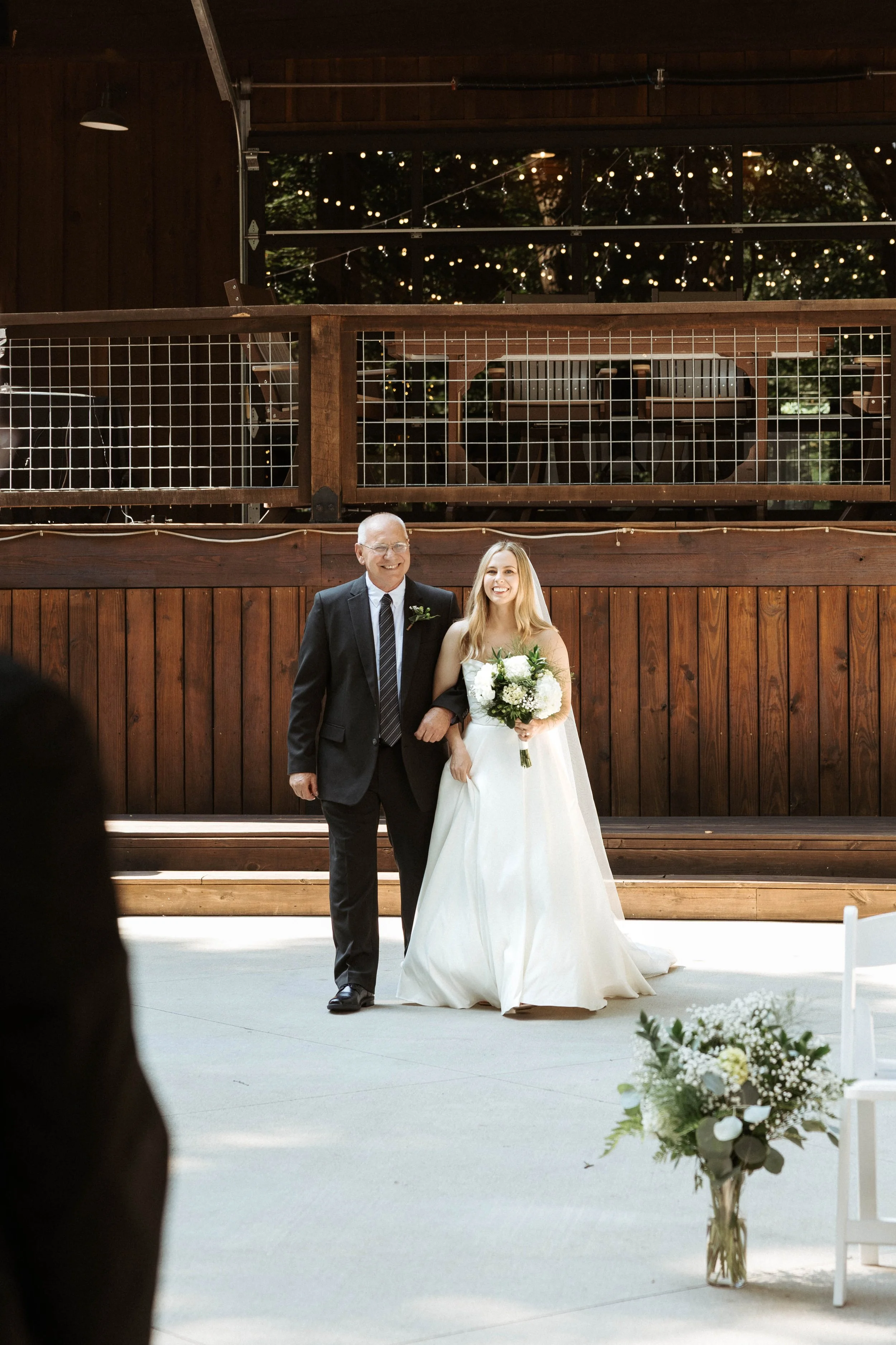 A bride in a white wedding dress walking down the aisle with an older man in a black suit, holding her arm, at a wedding ceremony inside a wooden venue with string lights overhead.