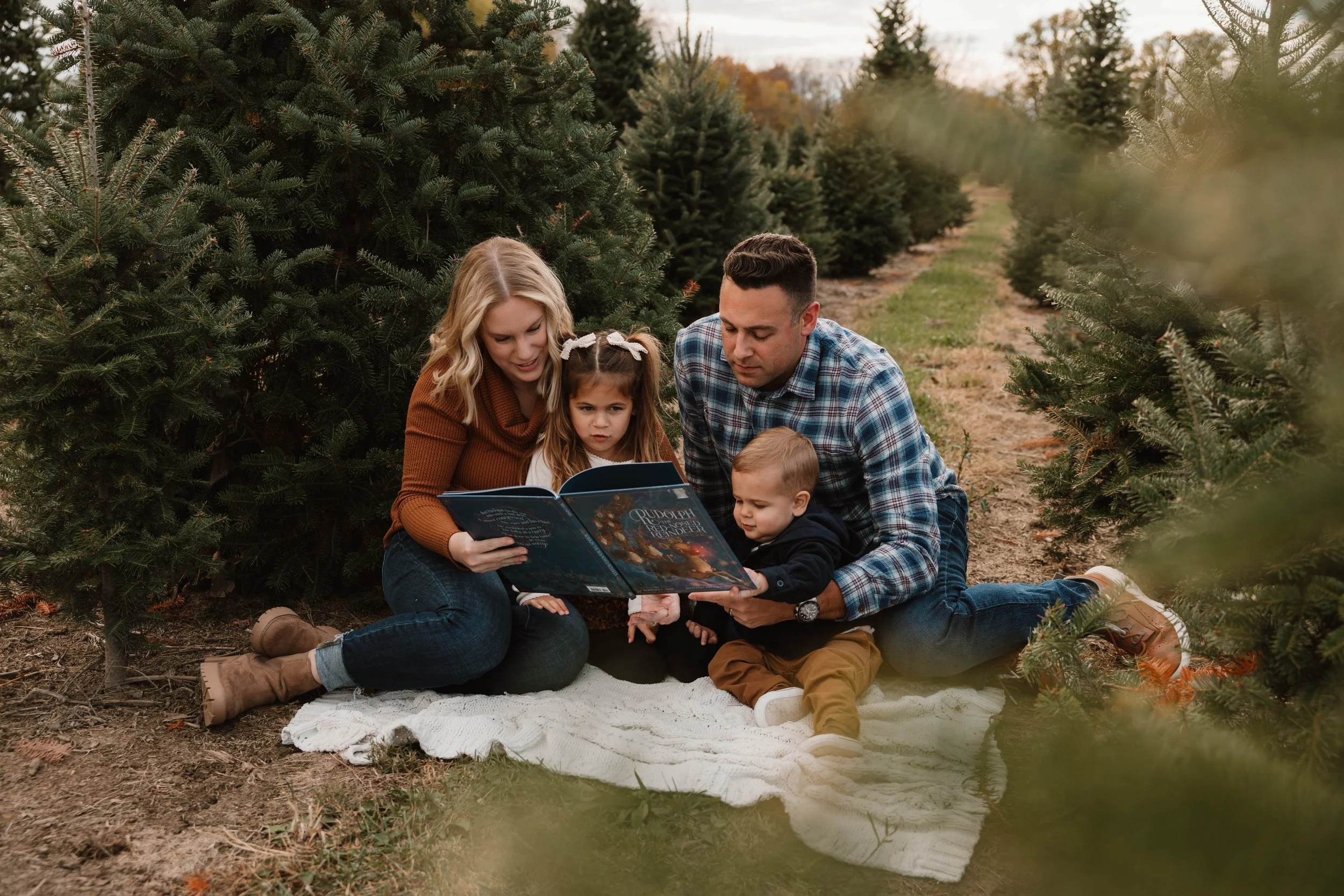 A family of five, including two children, sitting on a white blanket in a Christmas tree farm, reading a book together among evergreen trees during autumn.