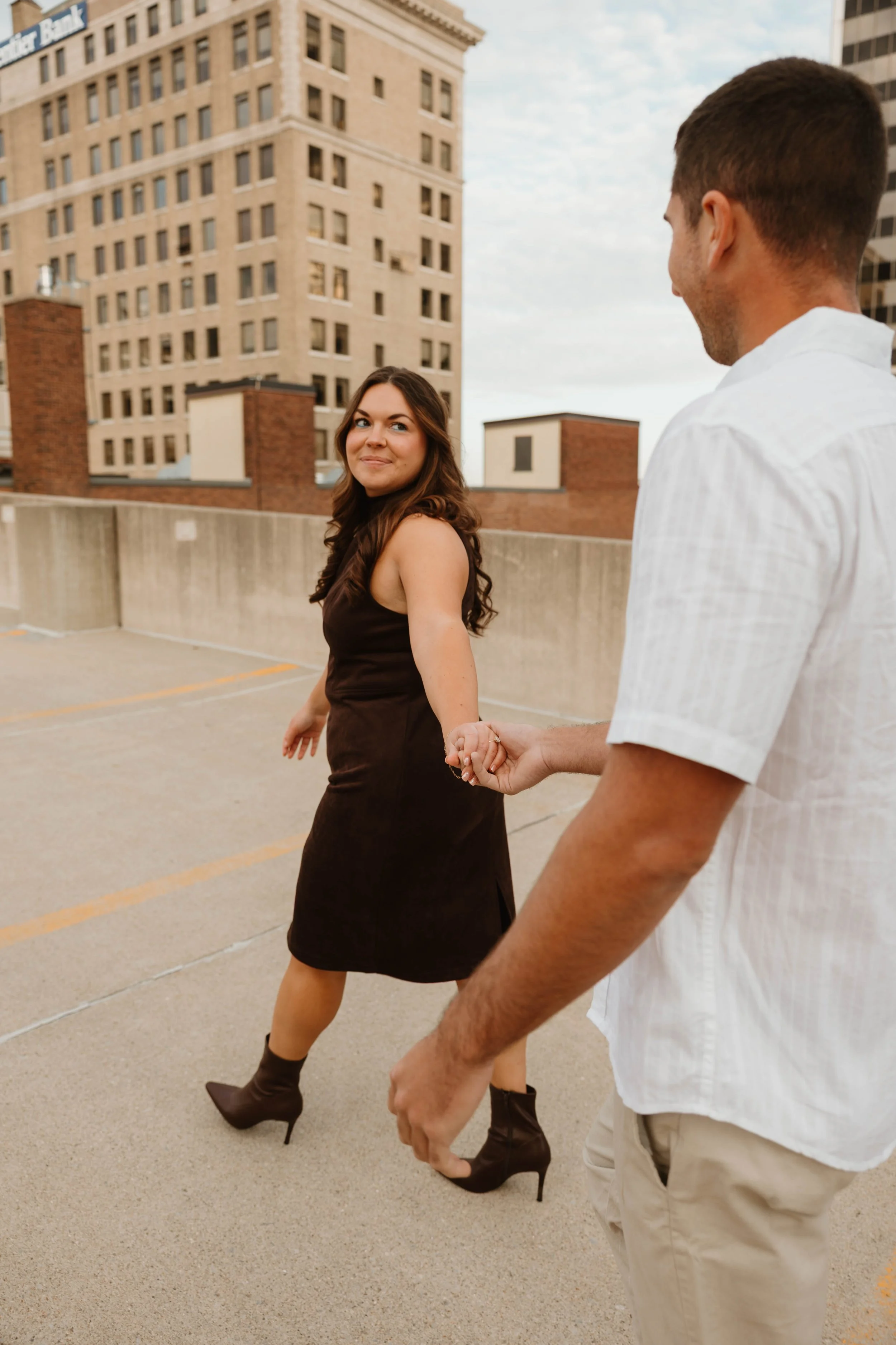 A man and woman holding hands on a rooftop parking lot with tall buildings in the background.