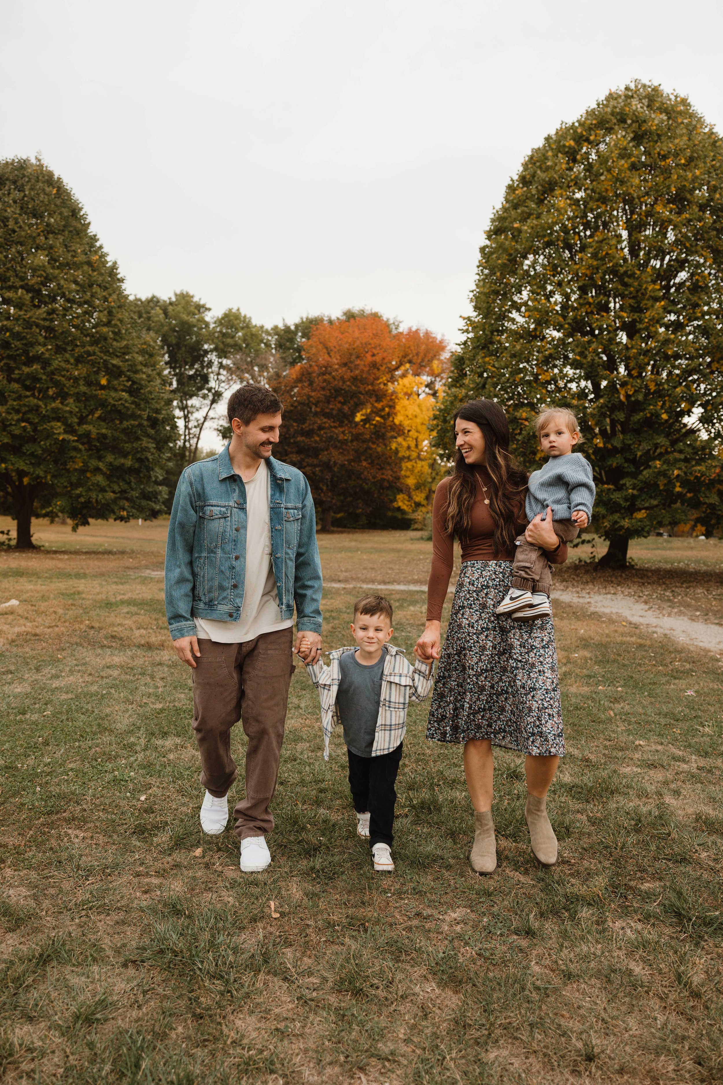 A family of four walking in a park with autumn trees in the background, holding hands and smiling.