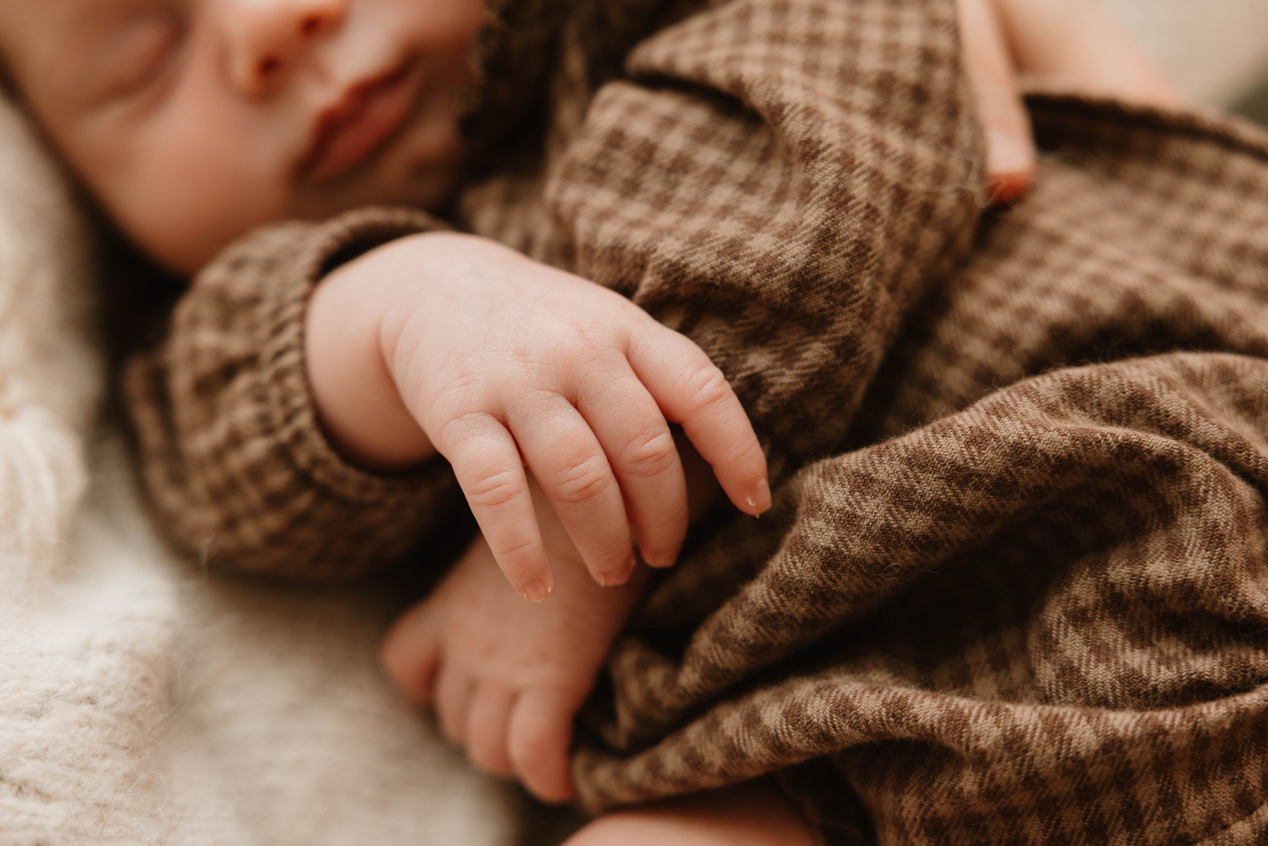 Close-up of a sleeping baby holding an adult's finger, wearing brown checkered clothing.