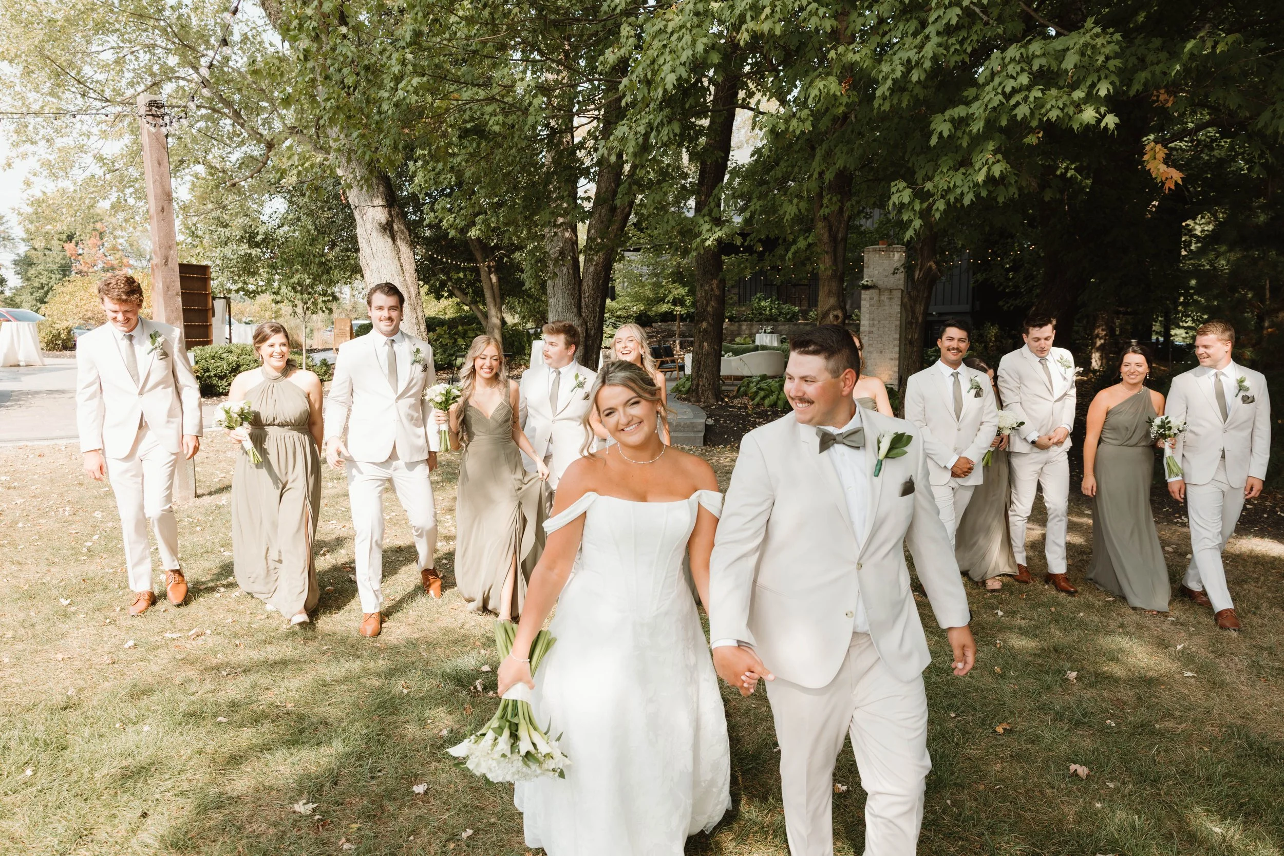 A wedding party walking outdoors on a sunny day, including a bride in a white dress holding a bouquet, a groom in a light-colored suit, and bridesmaids and groomsmen in matching dresses and suits, surrounded by trees.
