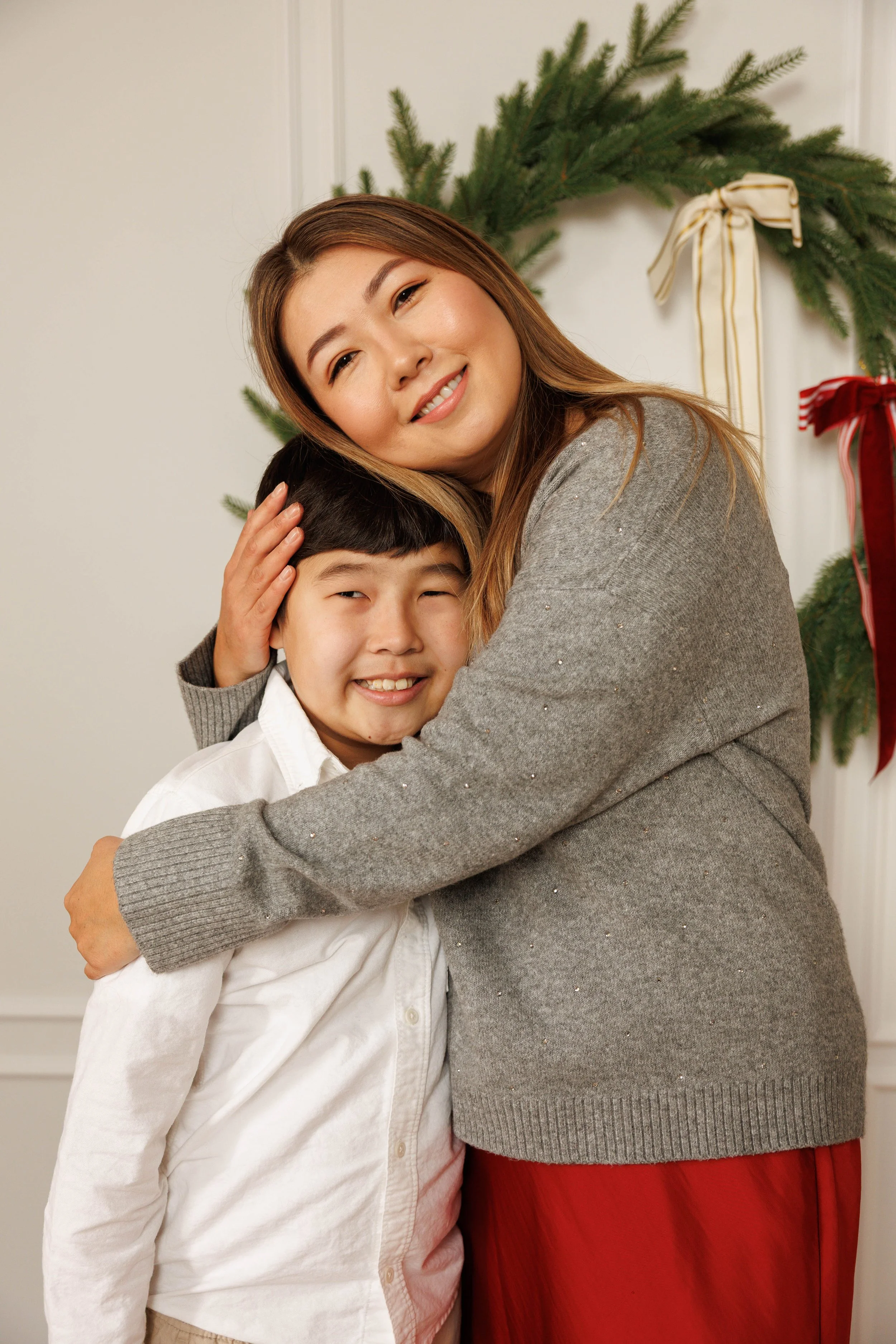 A woman and a young boy hugging in front of a Christmas wreath decorated with bows.