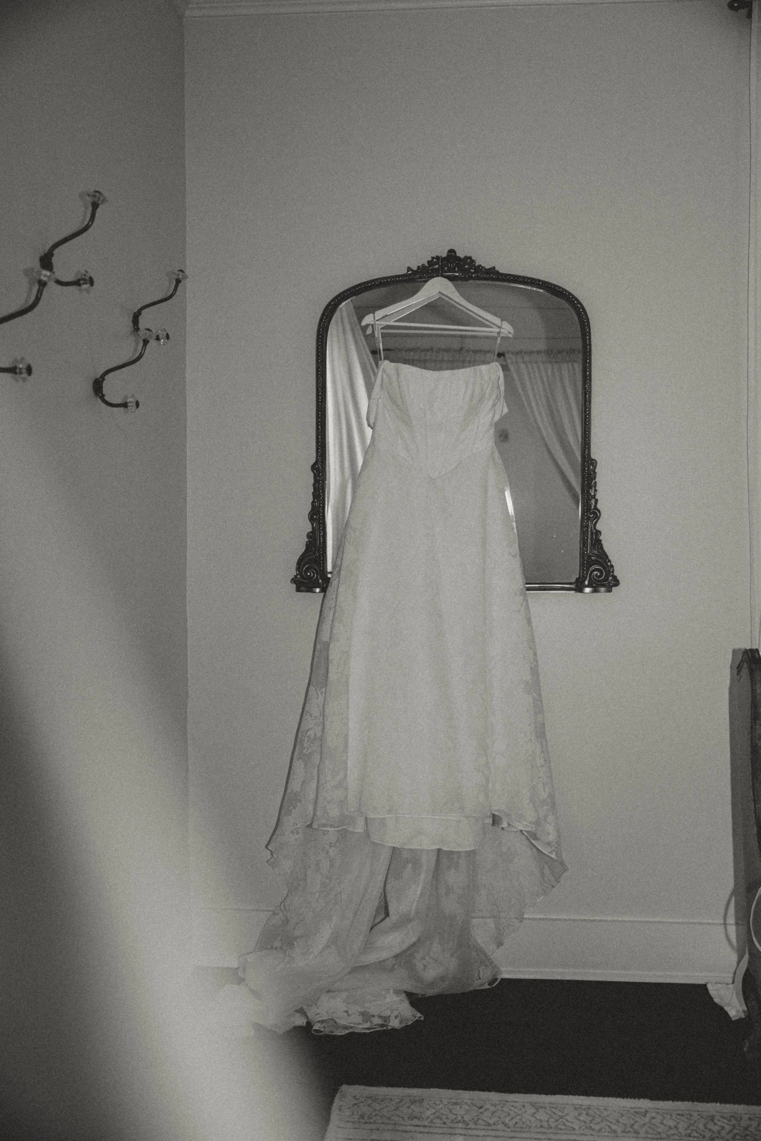 A wedding dress hanging on a hanger from a decorative mirror, with curtains and a partial view of a chair in the background.