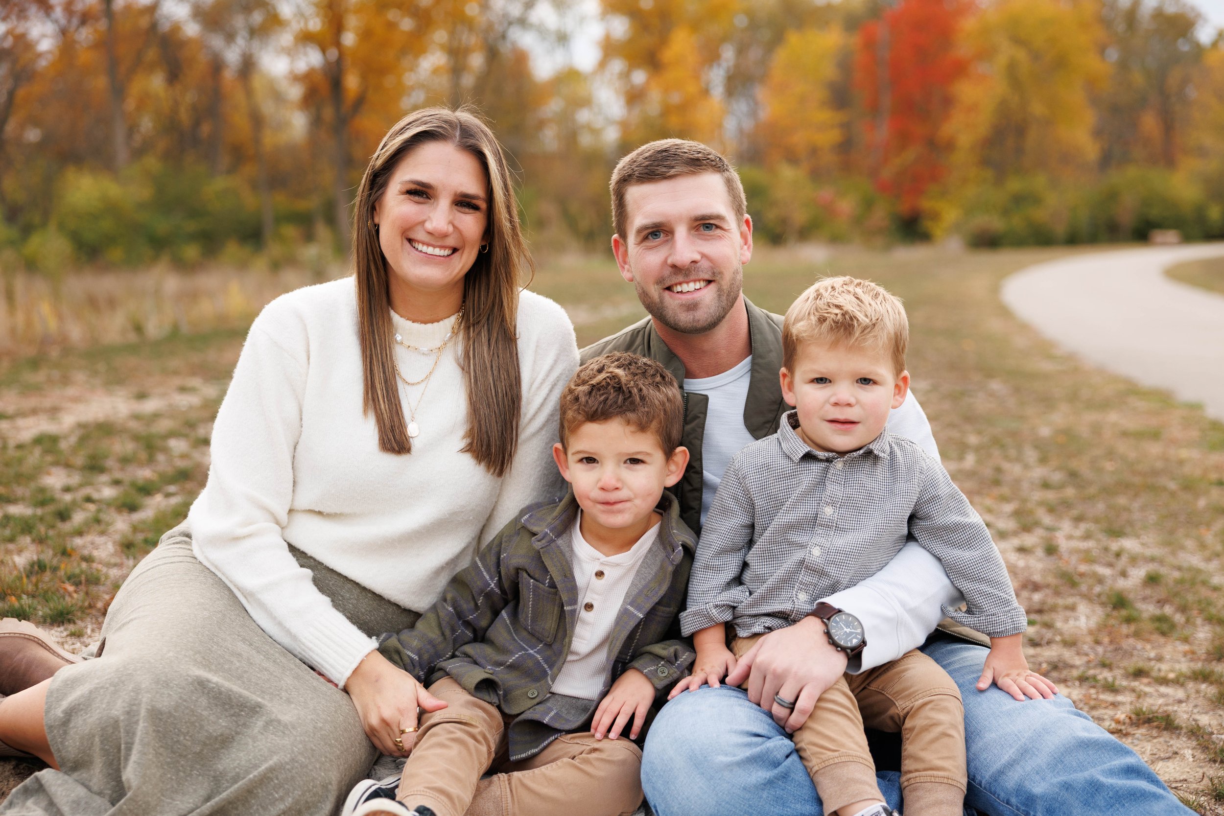 A happy family of five sitting outdoors on a fall day with colorful trees in the background. The mother and father are seated with their three young boys. The family is smiling and dressed in casual clothes.