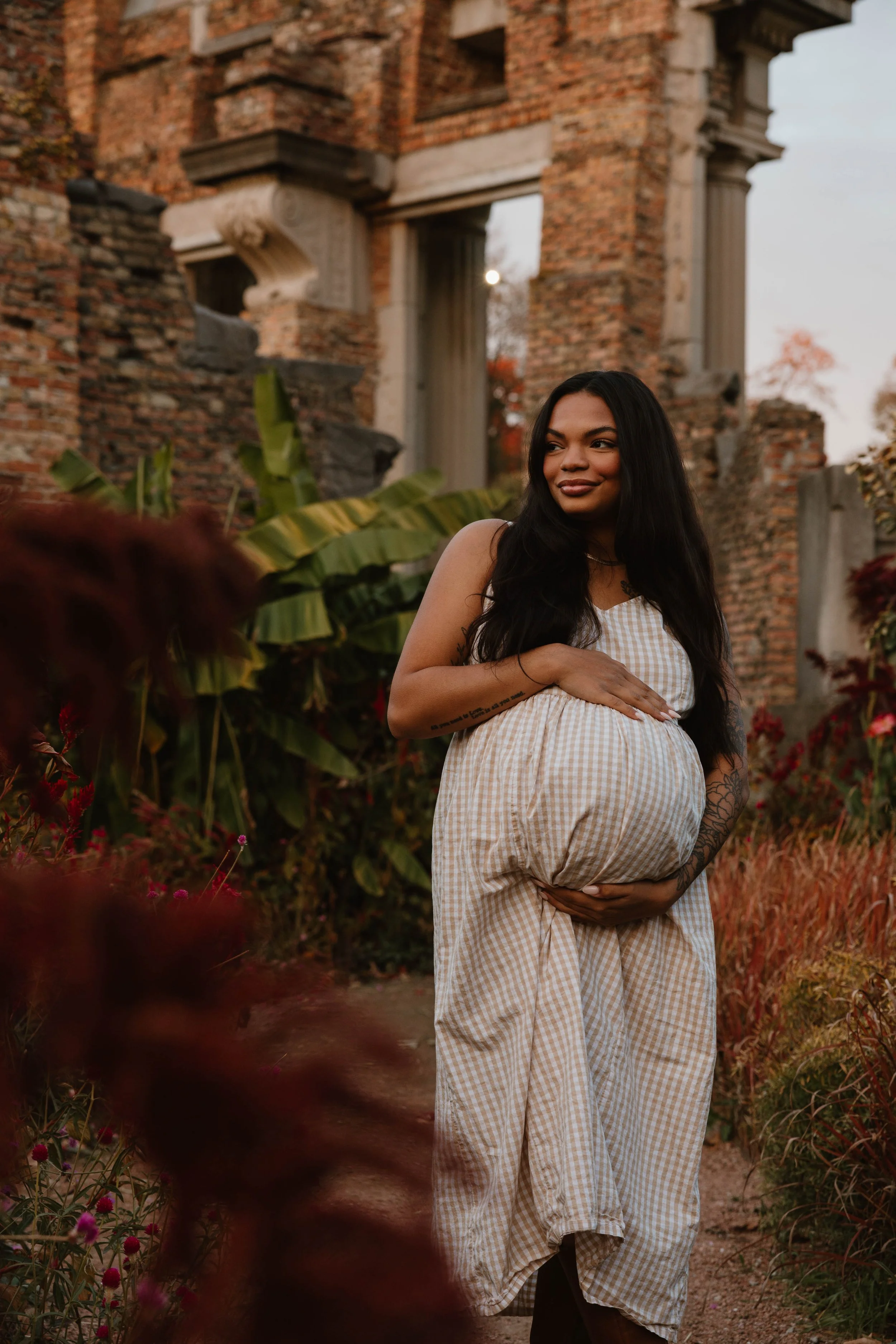 A pregnant woman standing in a garden with greenery and flowers, holding her belly, with ancient brick ruins in the background during sunset.