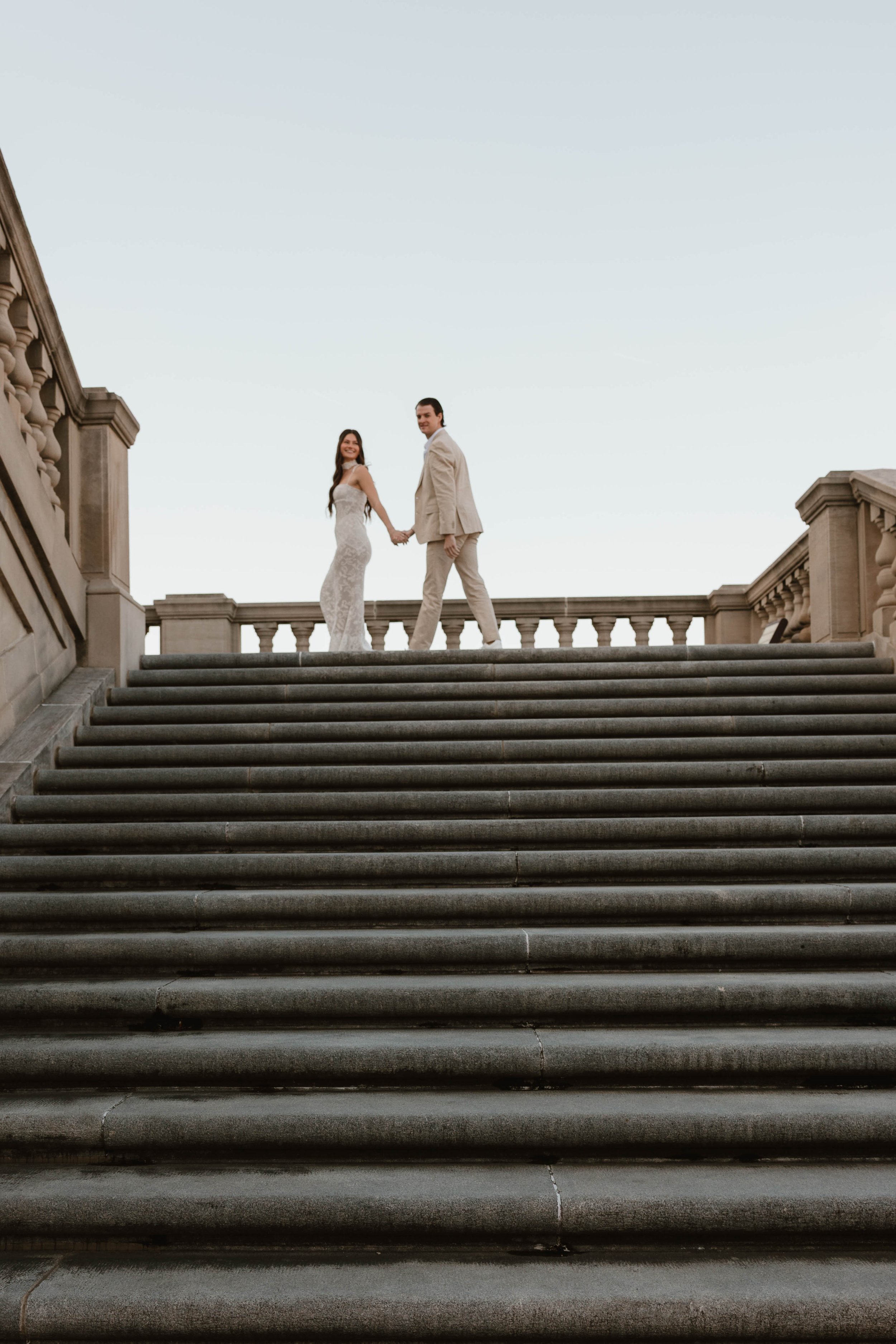 A bride and groom holding hands at the top of outdoor stone staircase with historic building railings, against a clear sky.
