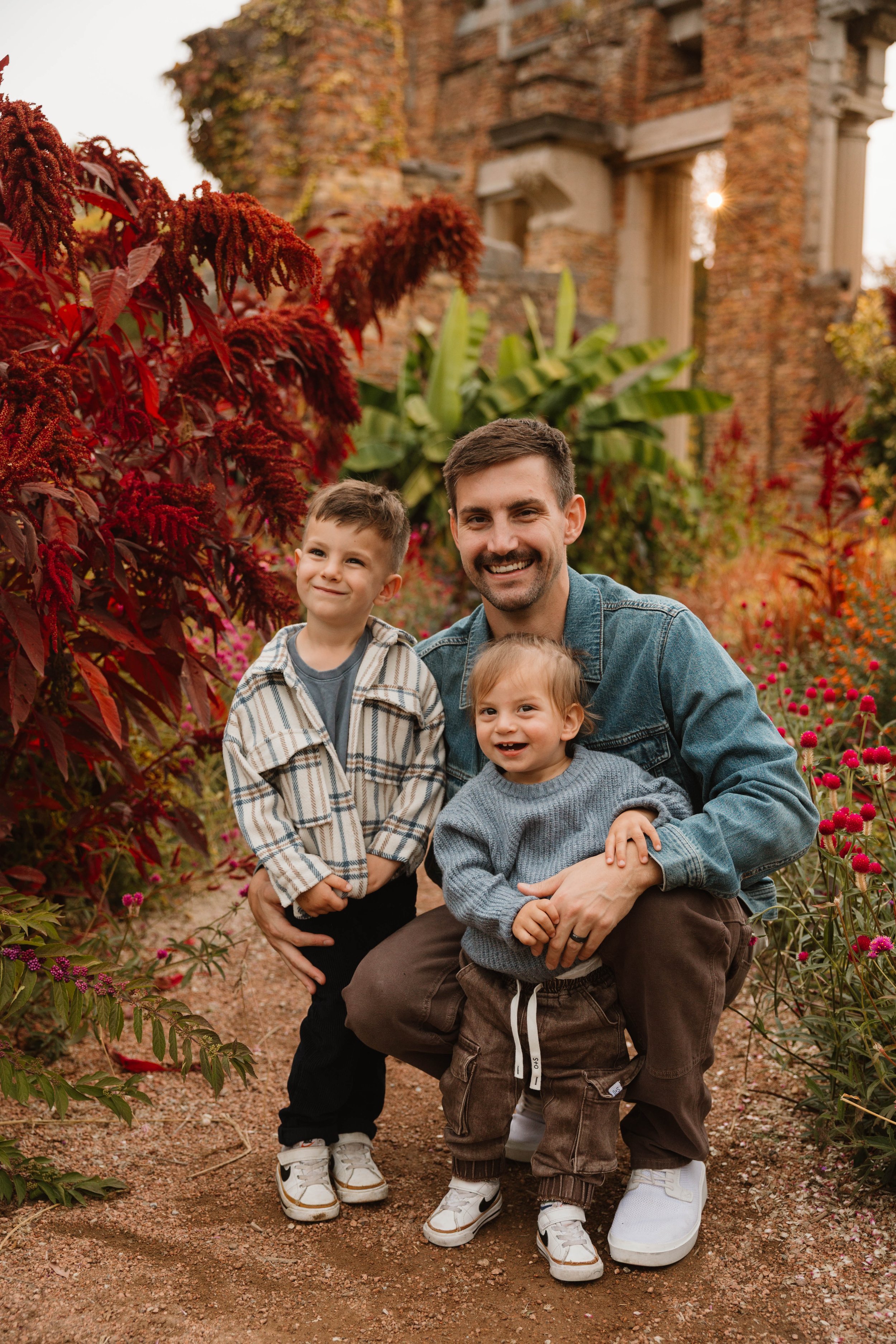 A man kneeling in a colorful garden with two young children, one standing and one sitting on his lap, surrounded by vibrant autumn plants and an old brick building in the background.