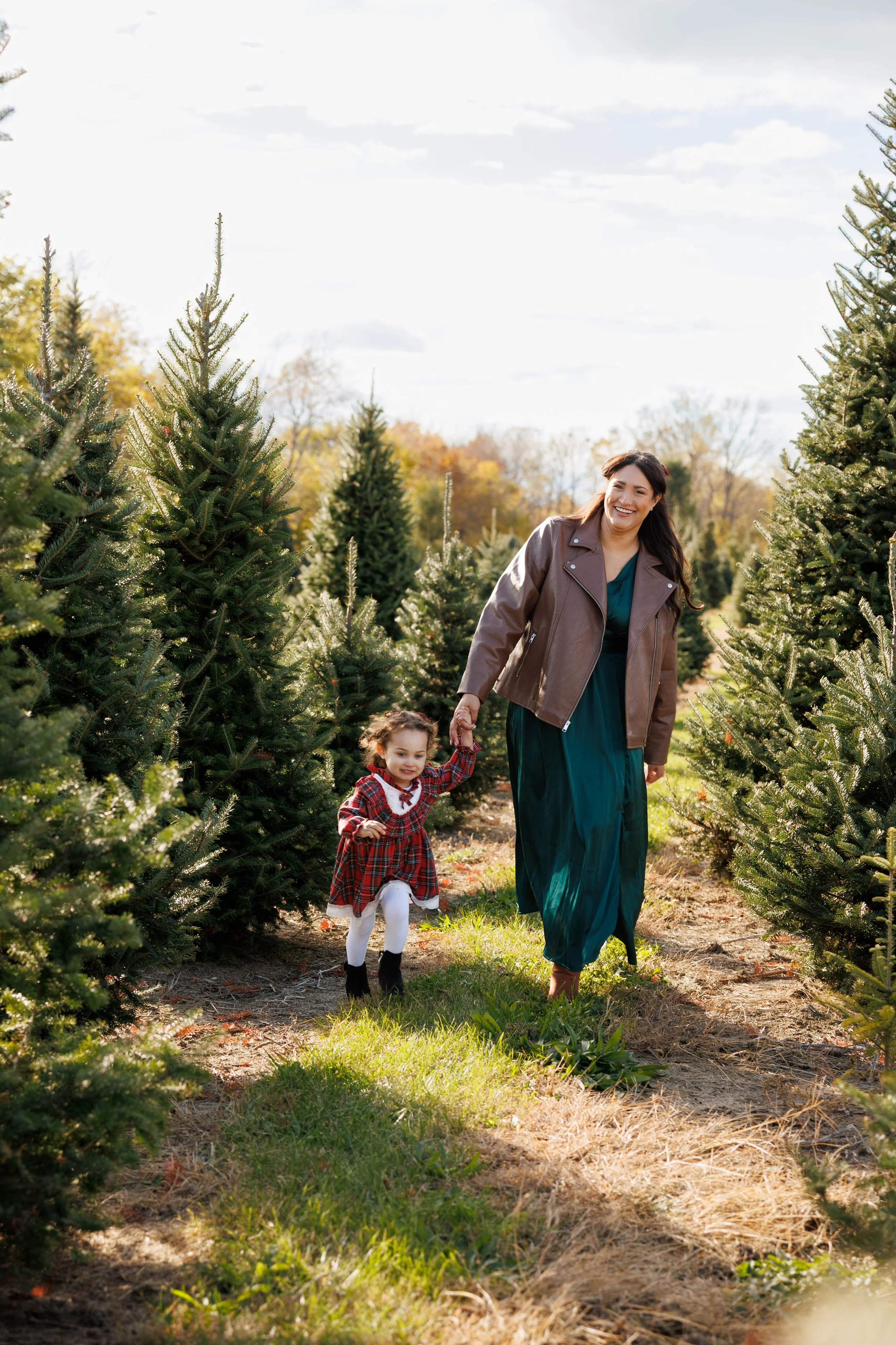 A woman and a young girl walking hand in hand through a Christmas tree farm with many evergreen trees, under a partly cloudy sky.