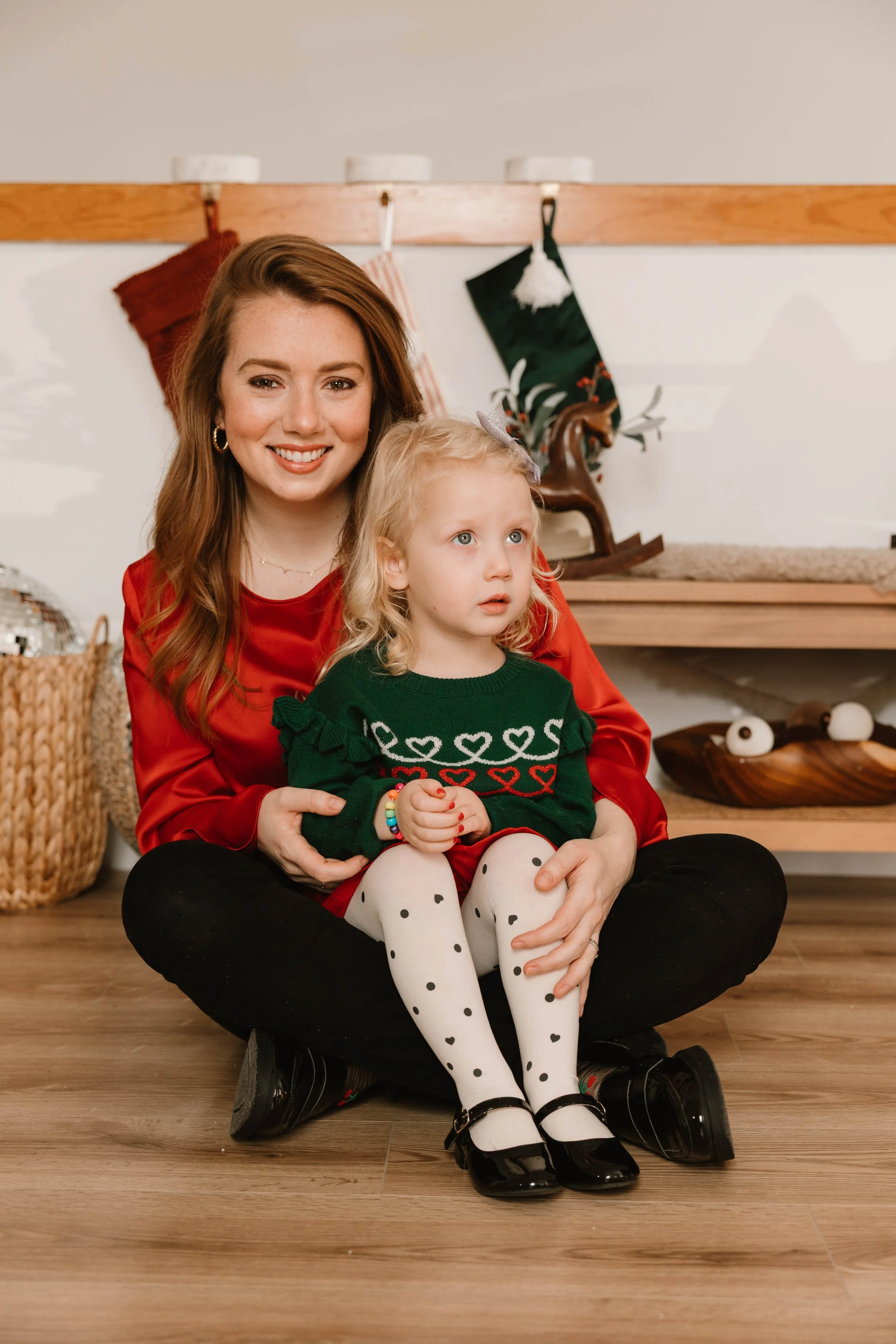 A woman and a young girl sitting on the wooden floor in front of a Christmas mantle. The woman is smiling and has long red hair, wearing a red top. The girl is looking away, wearing a green sweater with hearts, polka dot tights, and black shoes.