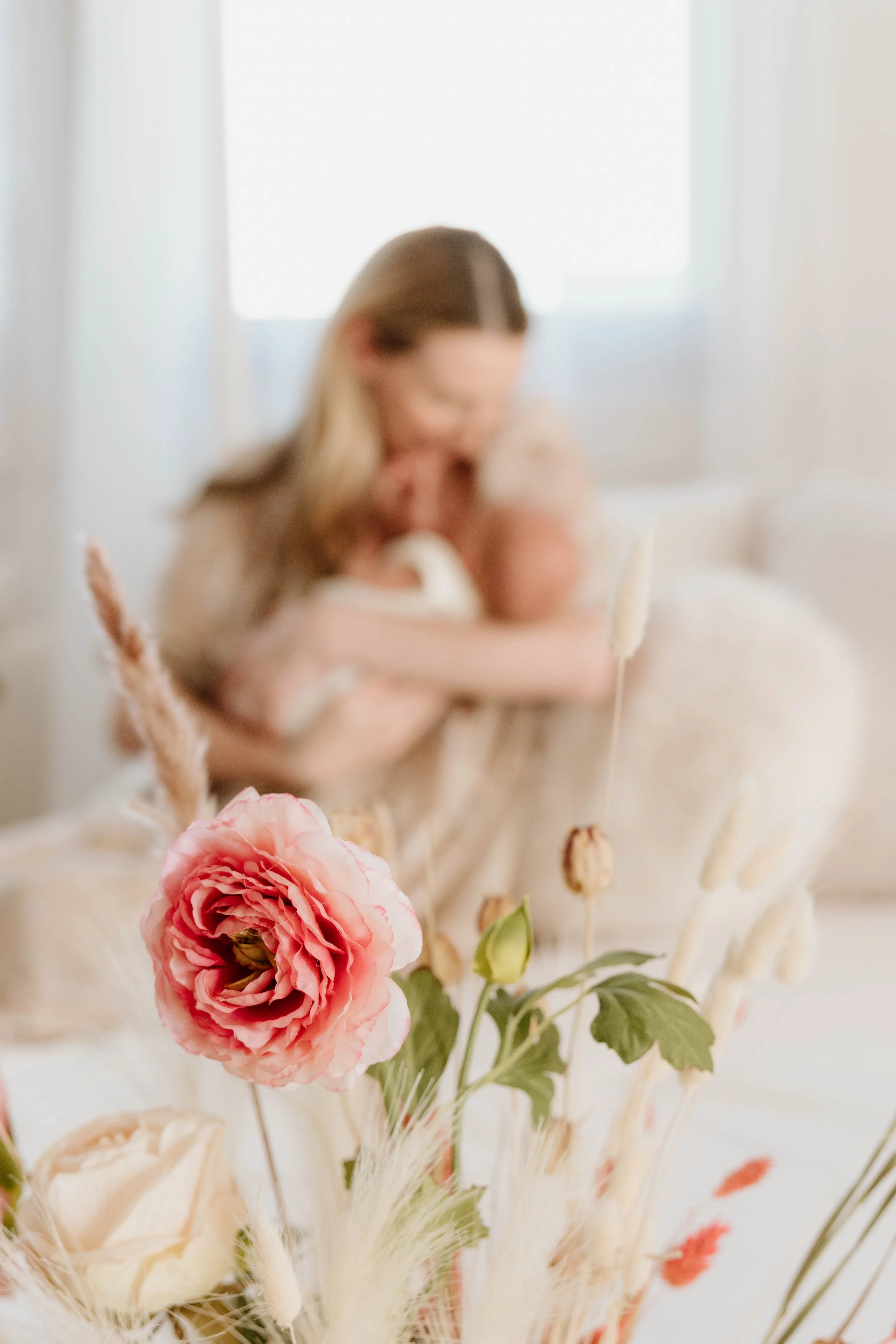 A woman embracing a baby in a bright, softly lit room with a bouquet of pink and white flowers in the foreground.