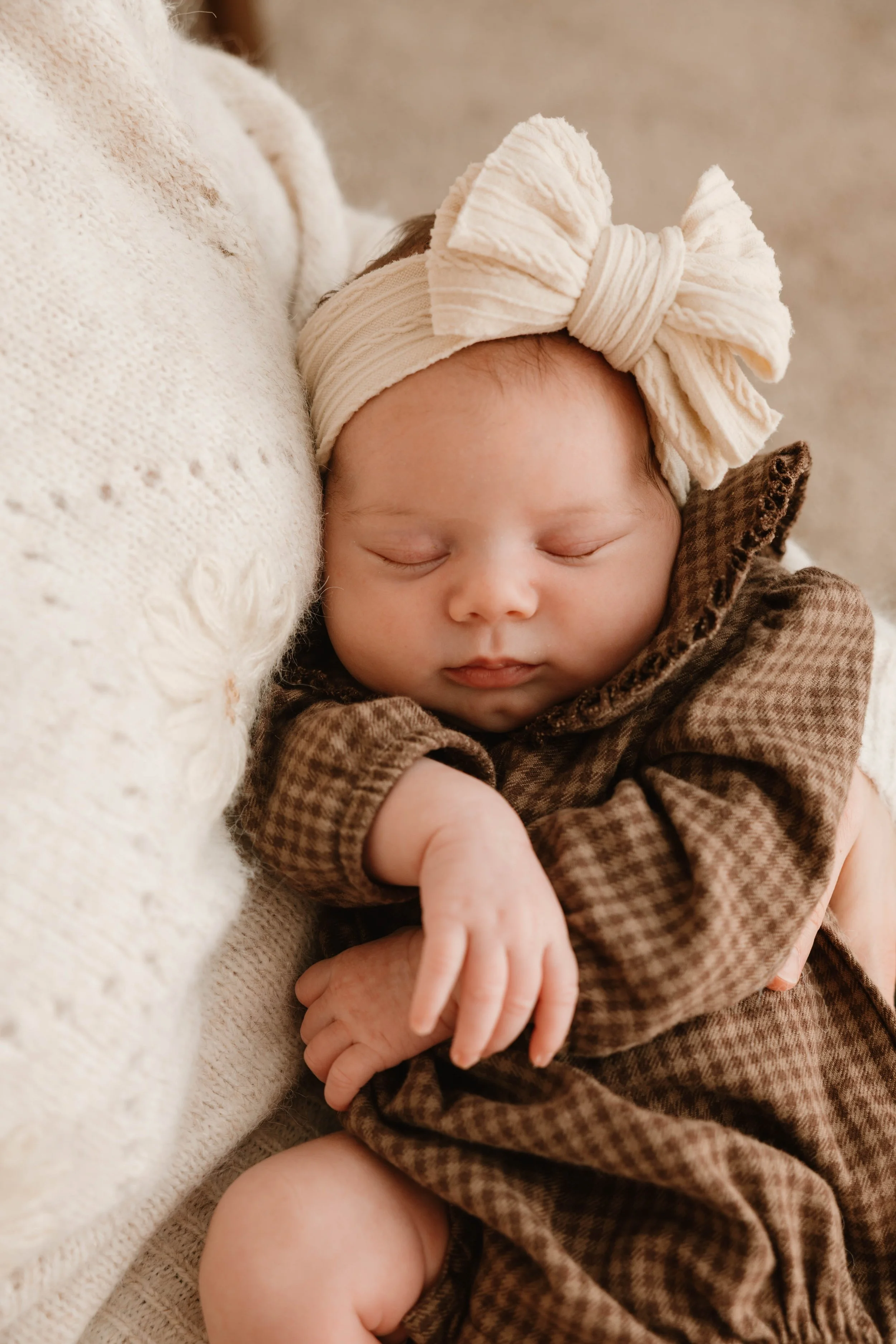 A sleeping baby with a large cream-colored headband with a bow, wrapped in a brown checkered outfit, resting on a soft knit blanket.