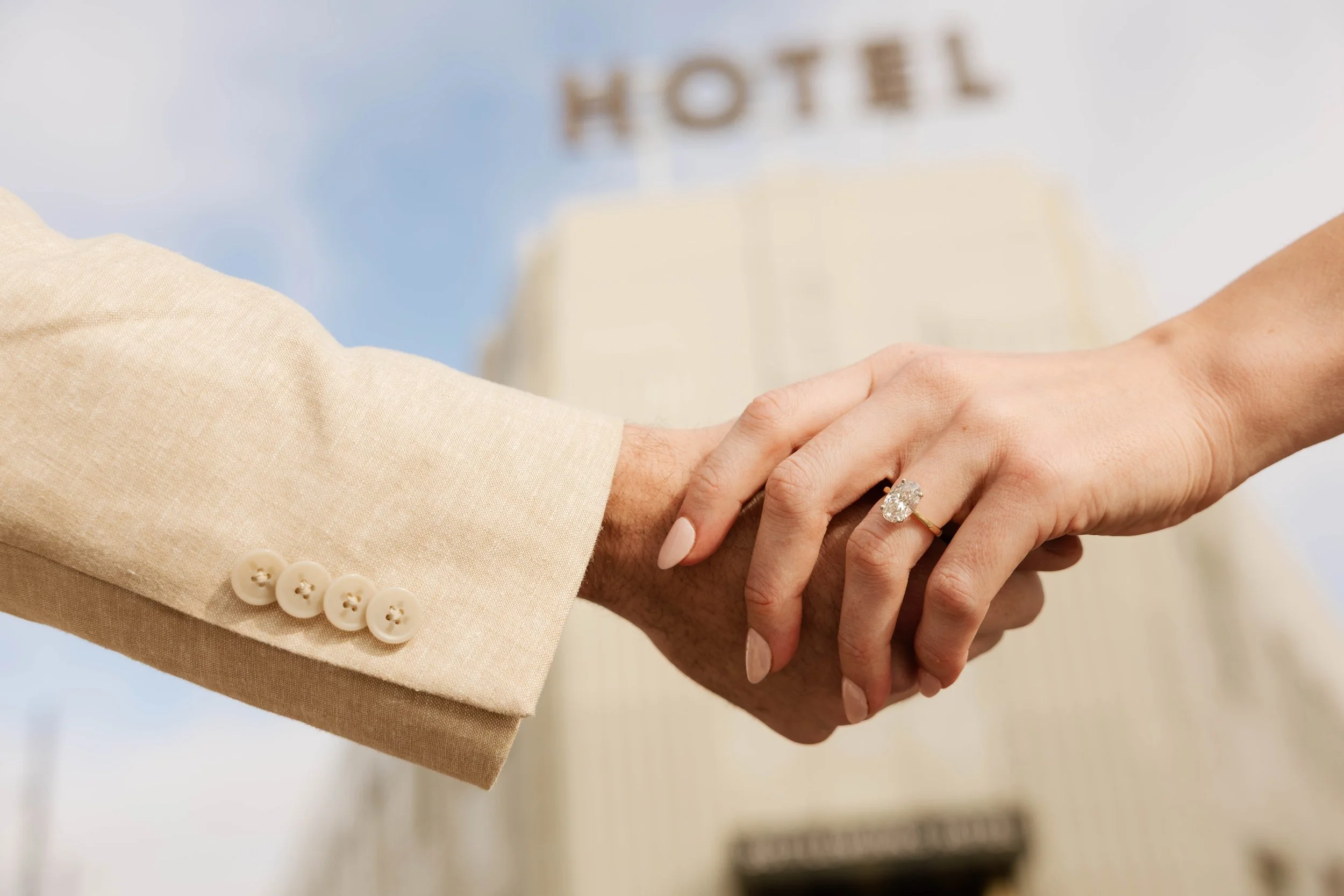 Close-up of a wedding handshake with a woman's hand displaying a diamond ring, and a man's hand in a beige suit, with a hotel building and blue sky in the background.