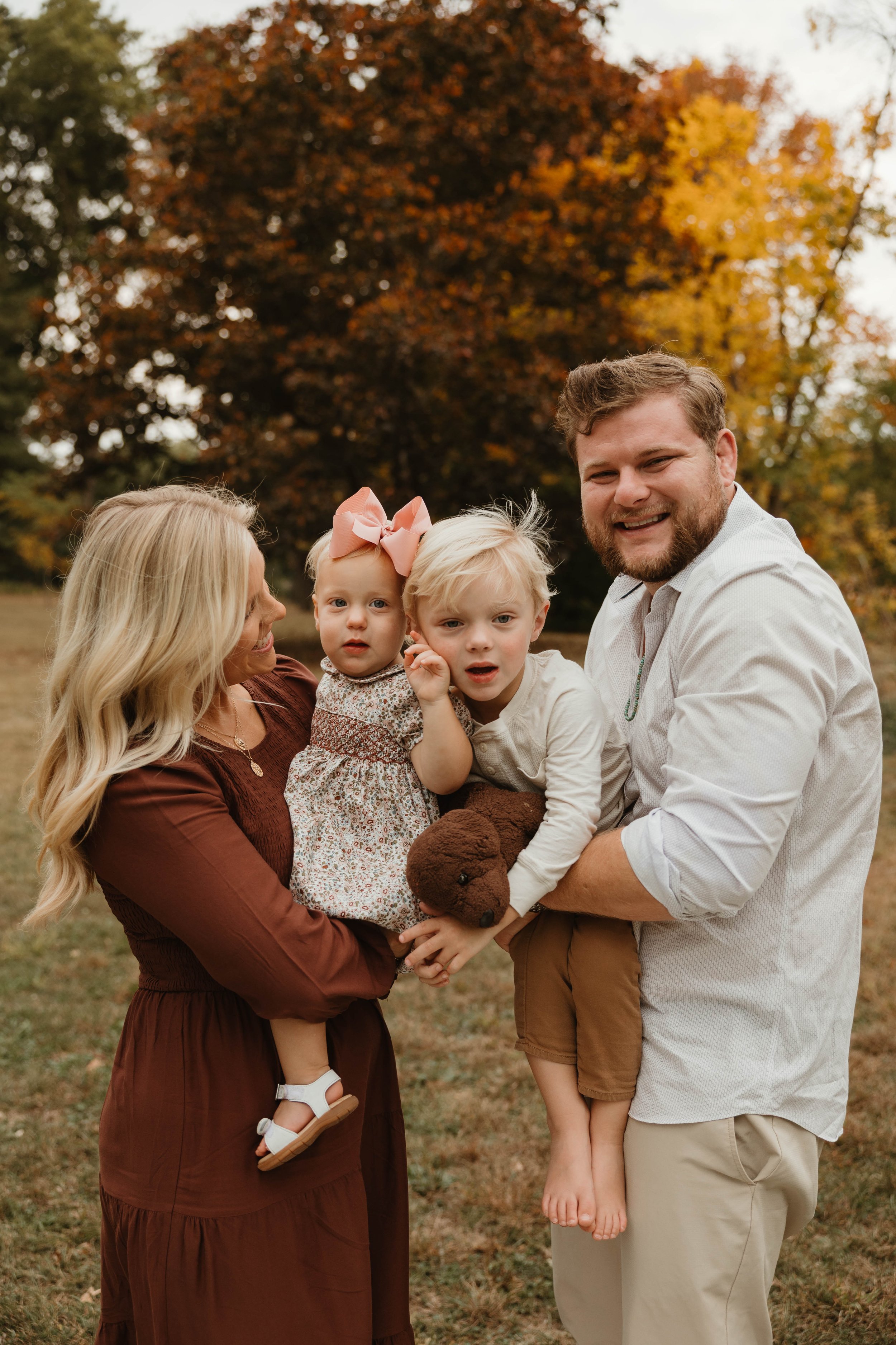 Family of four outdoors in fall, with colorful autumn trees in the background, smiling and holding their children and a plush teddy bear.