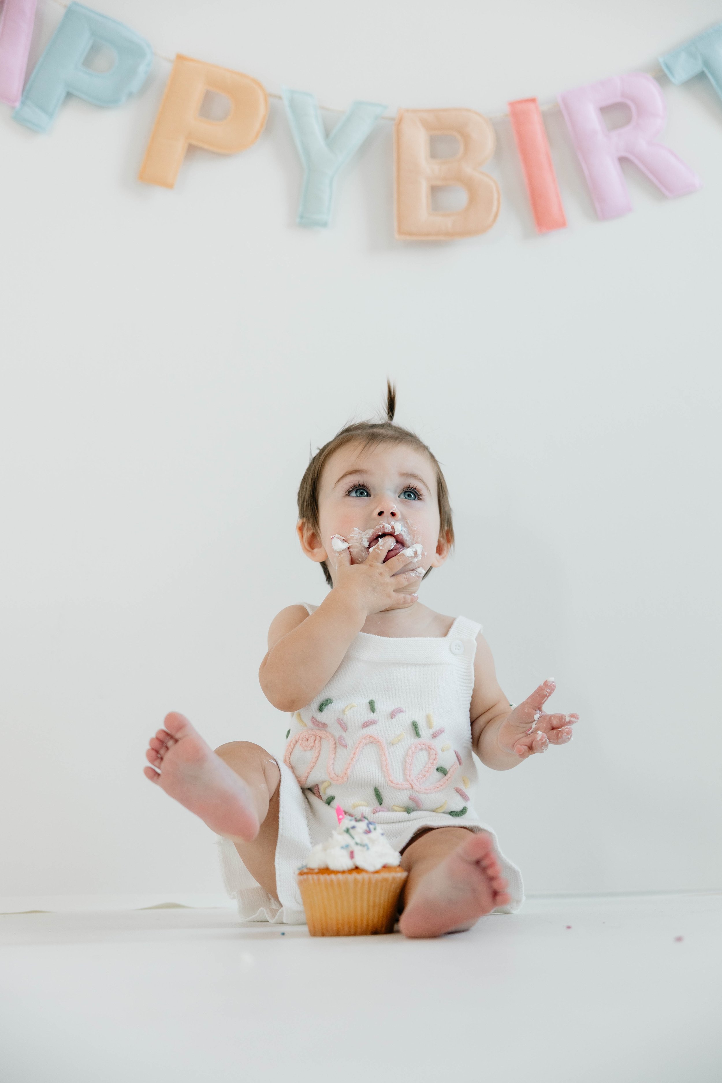 A young baby girl sitting on the floor celebrating her first birthday, with a colorful 'HAPPY BIRTHDAY' banner hanging above her and a cupcake in front of her.