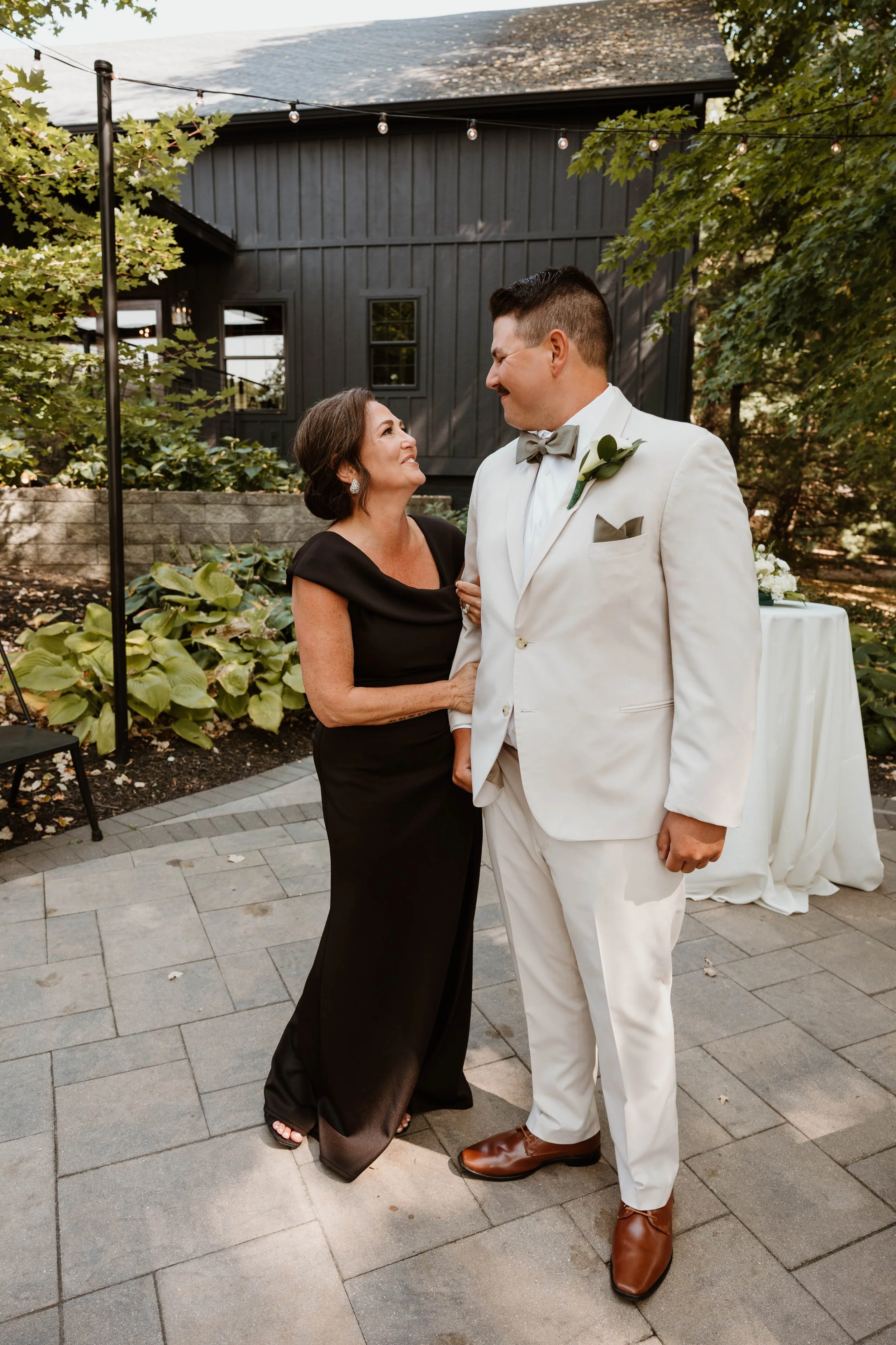 A woman in a black dress and a man in a white tuxedo with a grey bow tie and brown shoes are standing outside on a patio, smiling at each other. There are trees and string lights in the background.