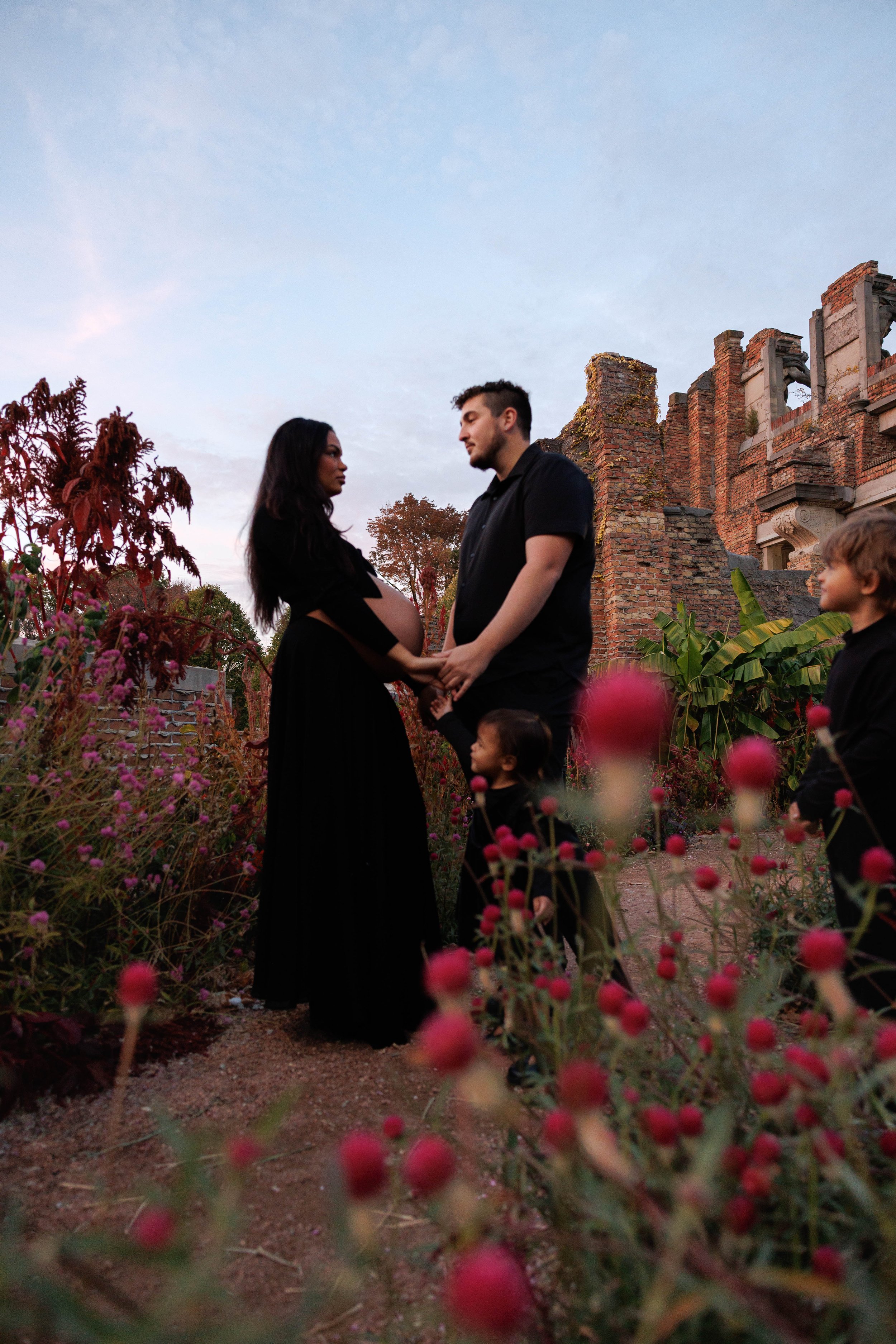 A couple stands holding hands in a garden with pink flowers, with two children nearby, and old brick ruins in the background during sunset.