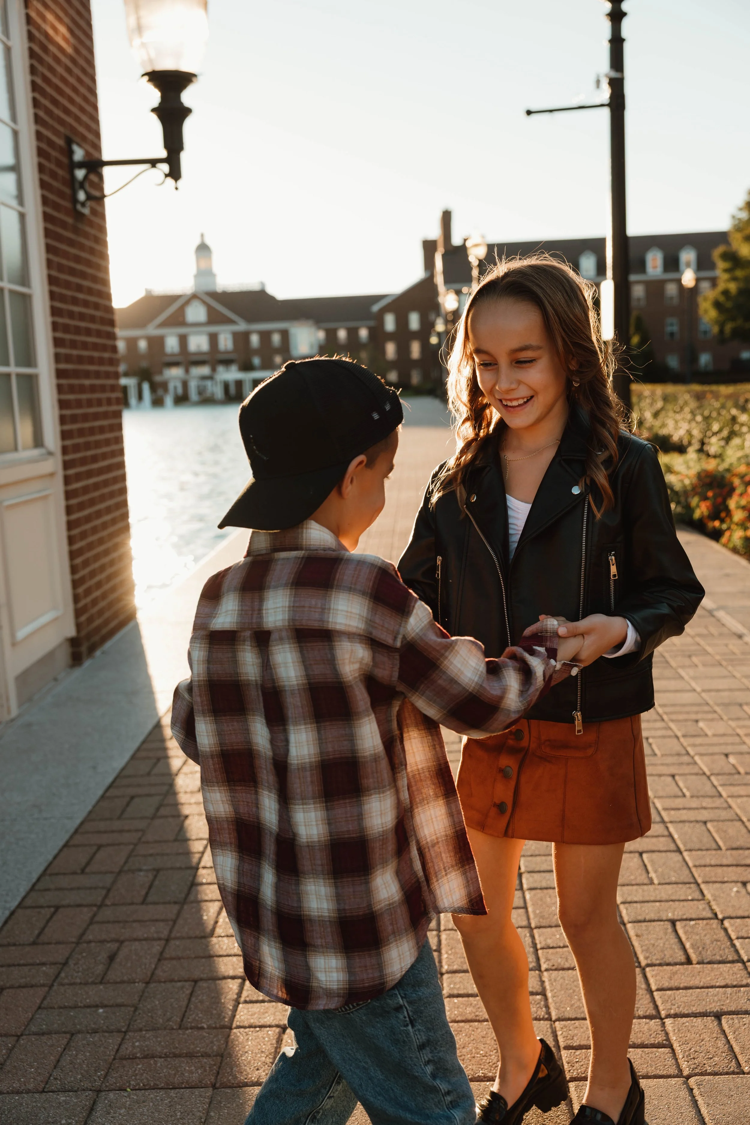 A young woman and a young boy are holding hands and smiling at each other outside near a body of water during sunset. The woman is wearing a black leather jacket and a brown skirt, and the boy is wearing a checkered shirt and a black cap.