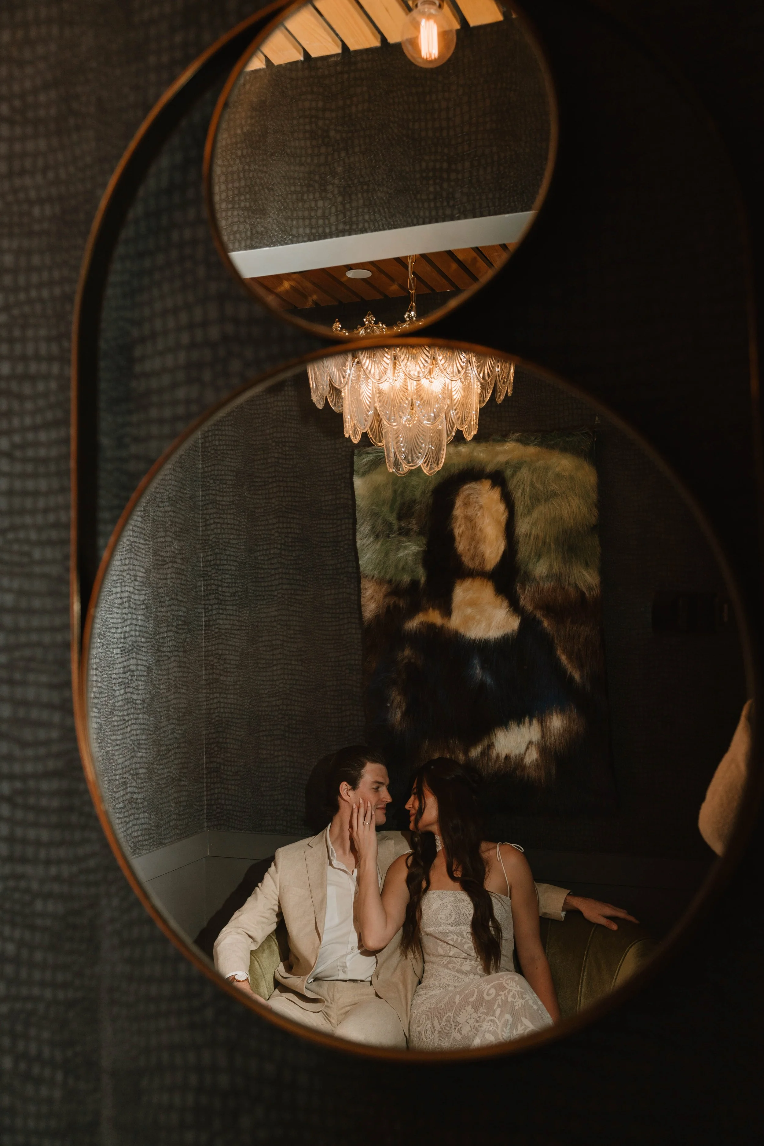 Couple sitting close together on a couch, facing each other, touching faces, in a dimly lit room with a chandelier and artwork in the background.
