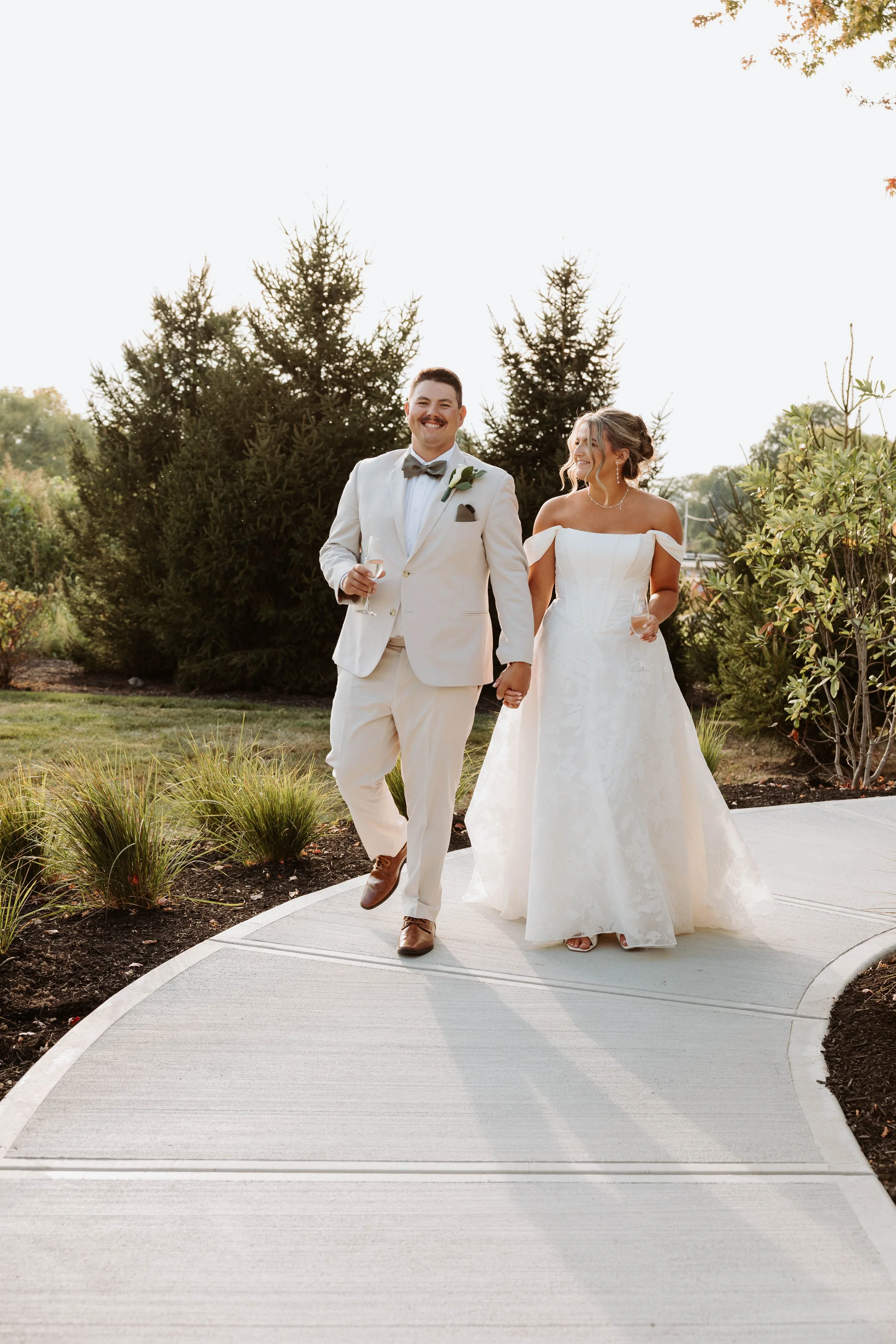 A couple in wedding attire walking outdoors on a curved pathway, holding hands, smiling, with greenery and trees in the background.