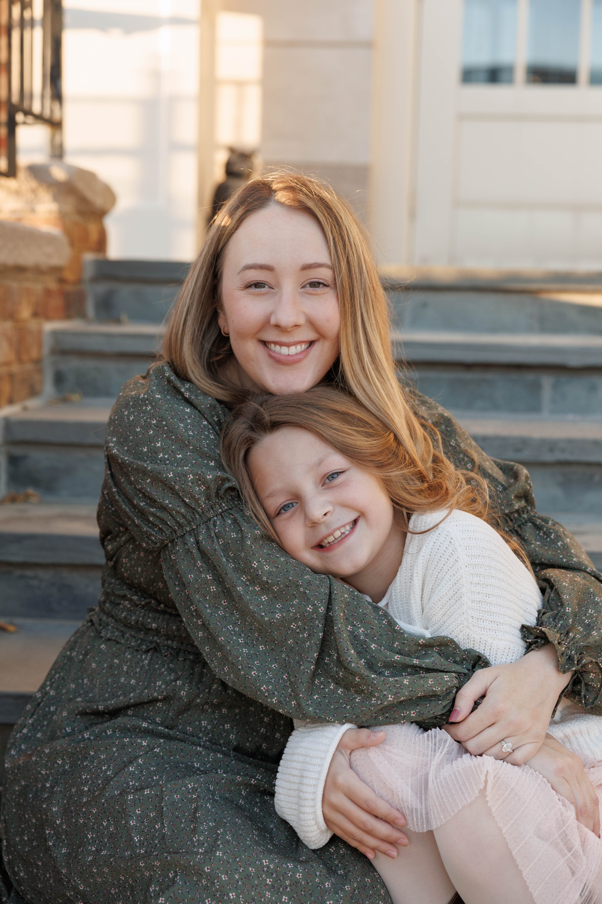 A woman and a girl sitting on outdoor steps, smiling and hugging each other.