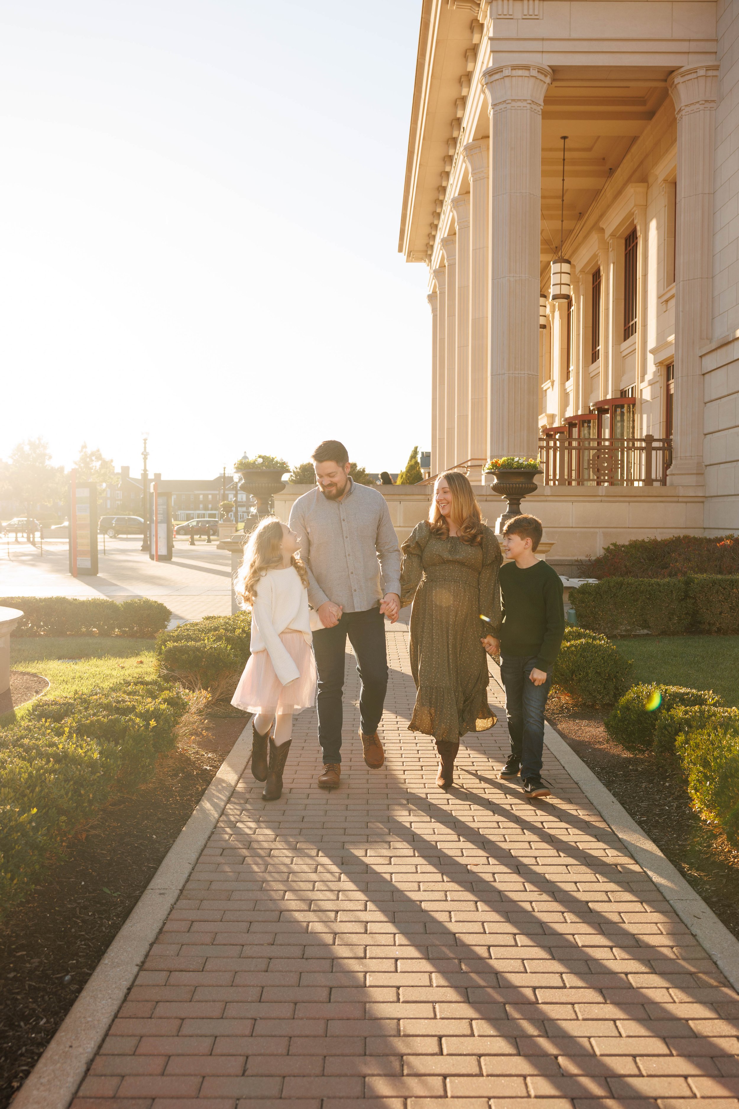 Family of four walking hand in hand on a brick pathway near a large building with columns, in warm sunlight.