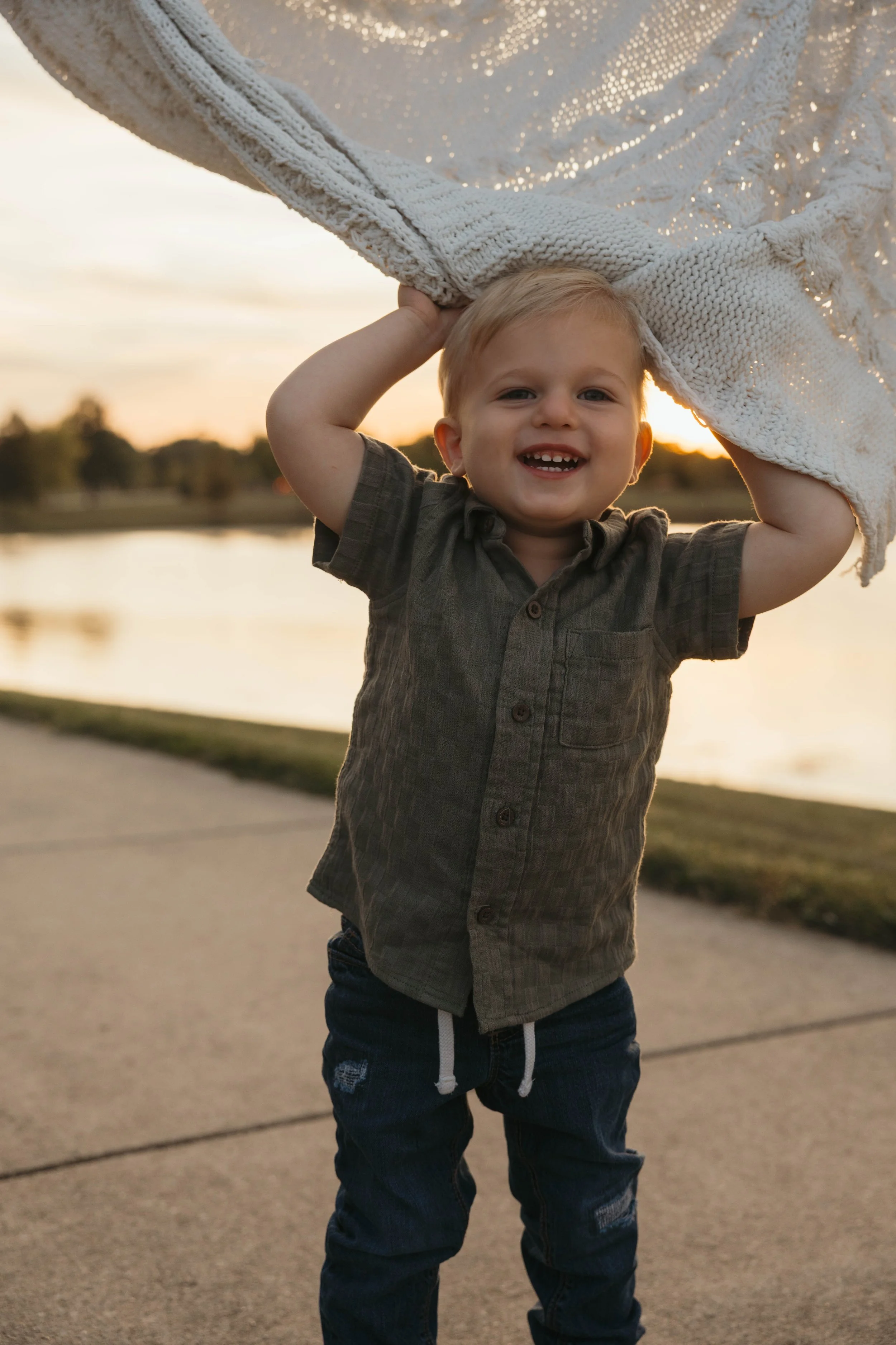 A smiling young boy holding a blanket over his head outdoors during sunset.