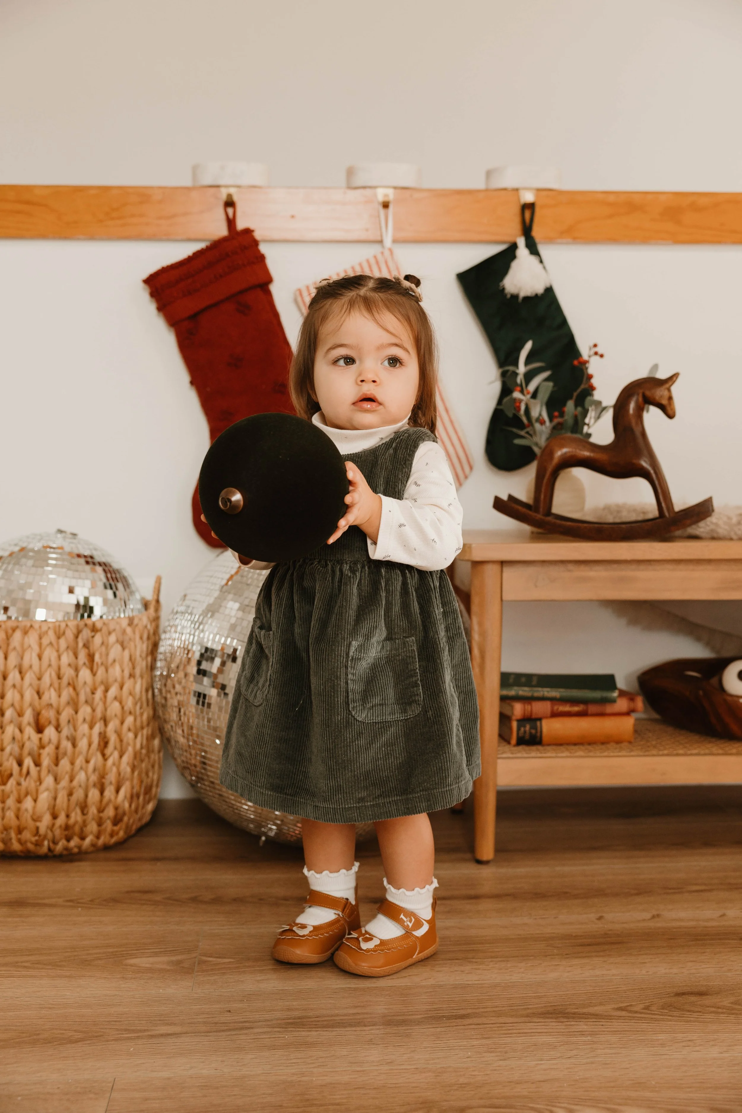 A young girl holding a black ball standing in a room decorated for Christmas, with stockings hanging on a wooden rail, a wooden rocking horse, and festive decor in the background.