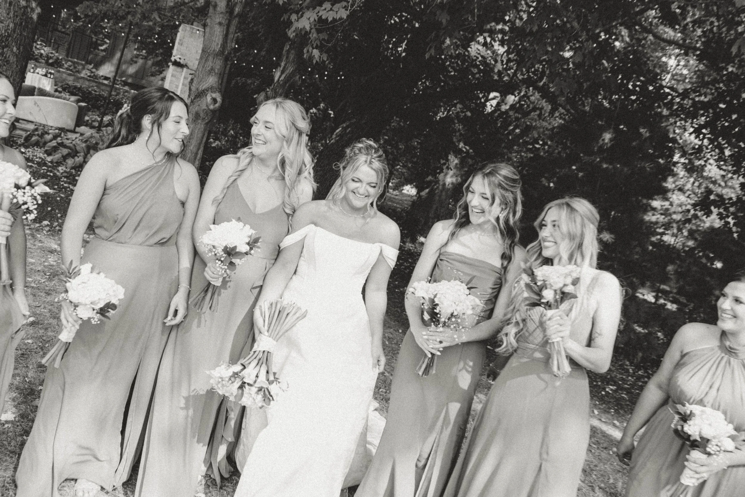 A bride and six bridesmaids standing outdoors, smiling and laughing, holding bouquets of flowers.
