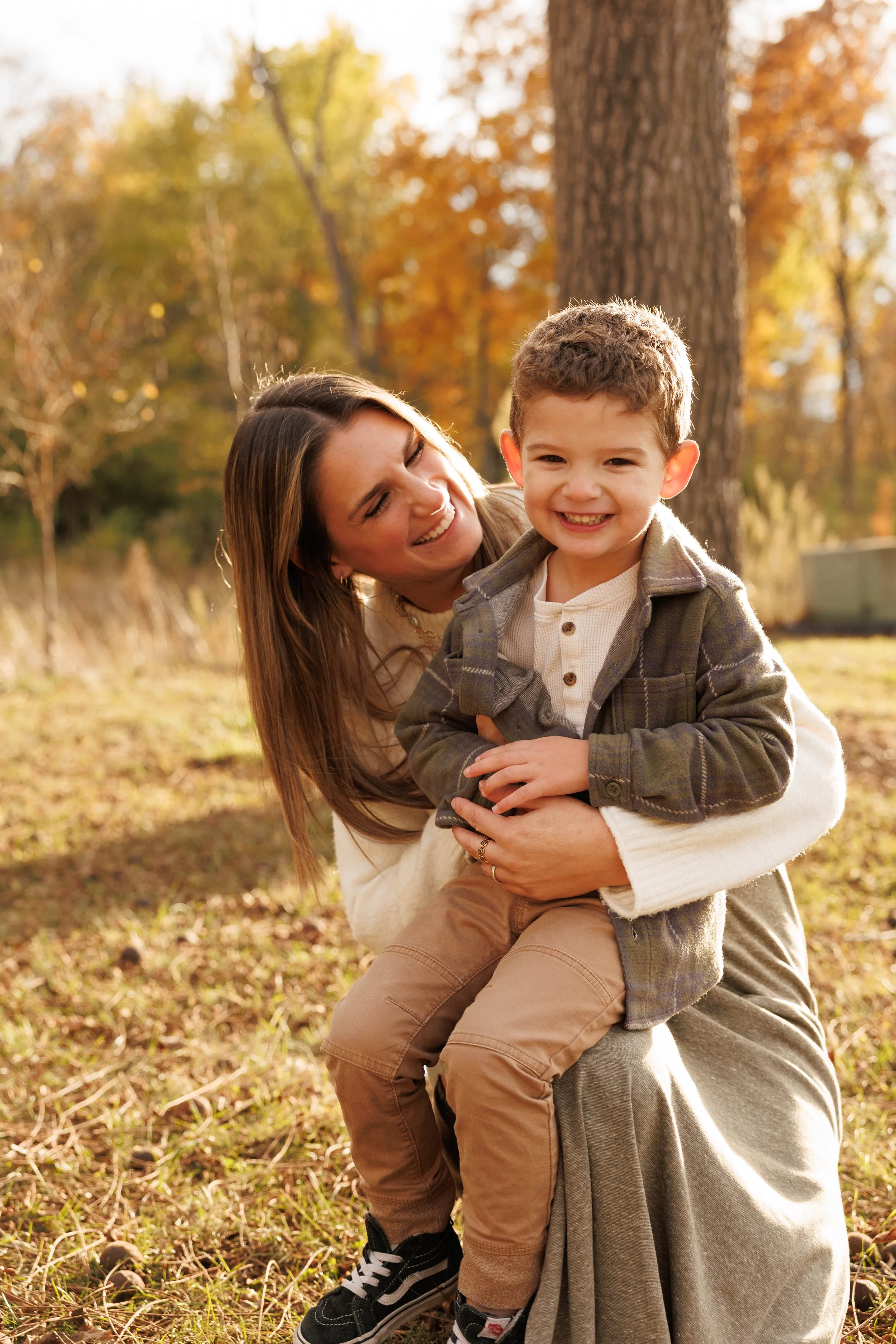 A woman and a young boy smiling and playing outside in autumn, surrounded by colorful fall trees and sunlight.