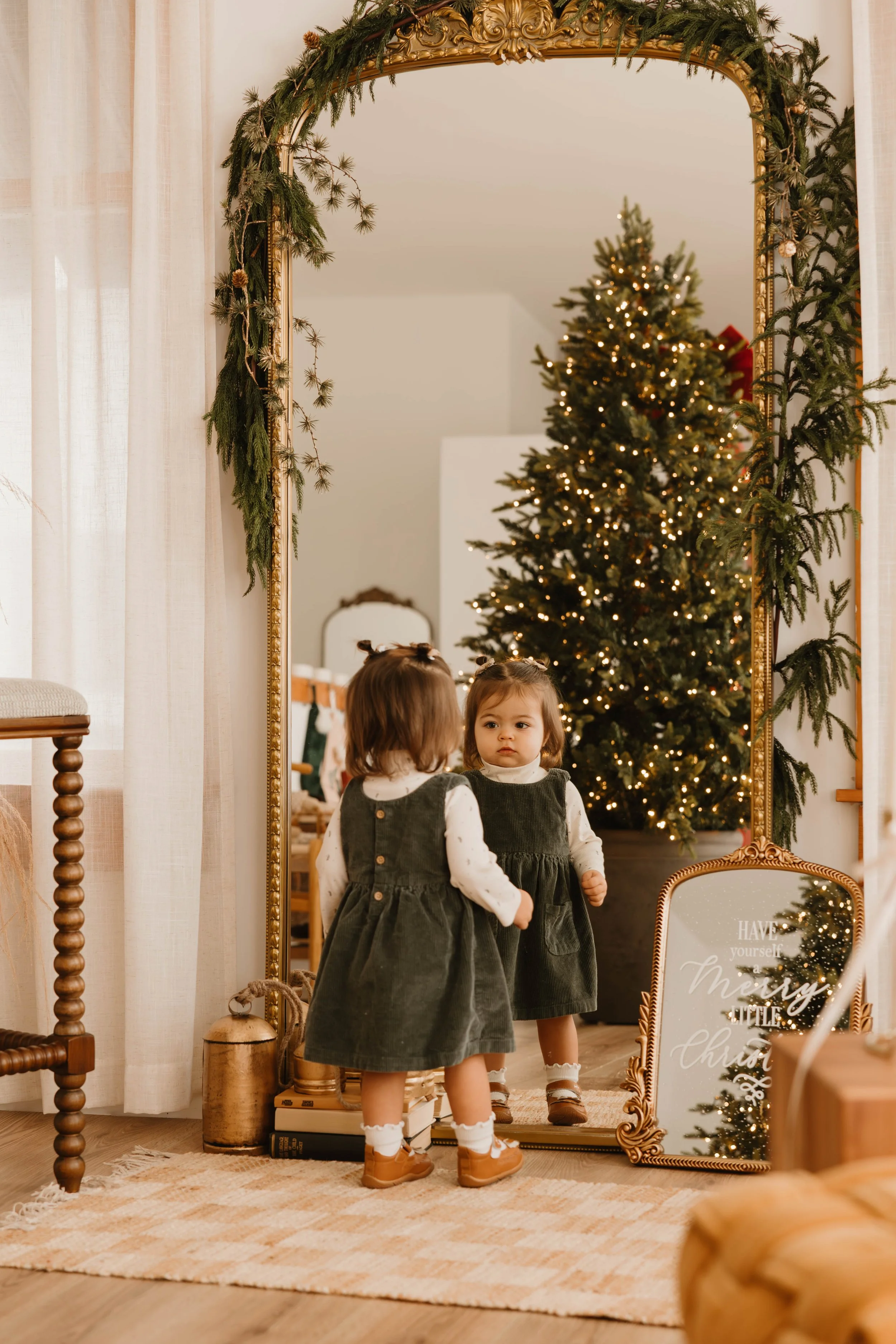 A young girl stands in front of a large mirror, looking at her reflection, with a decorated Christmas tree and festive decorations in the background.