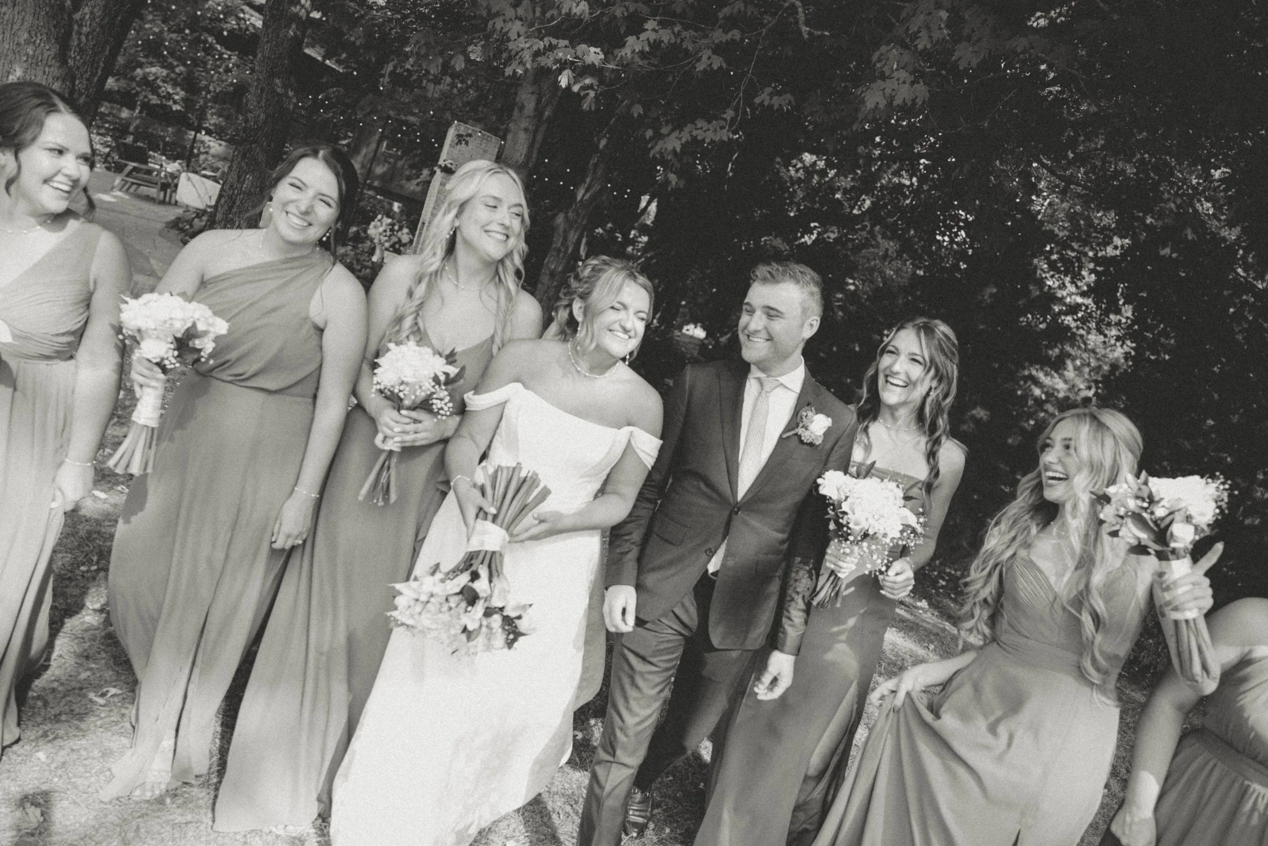 A black and white photo of a wedding party outdoors, featuring the bride, groom, and their bridesmaids and groomsmen, all smiling and holding bouquets.