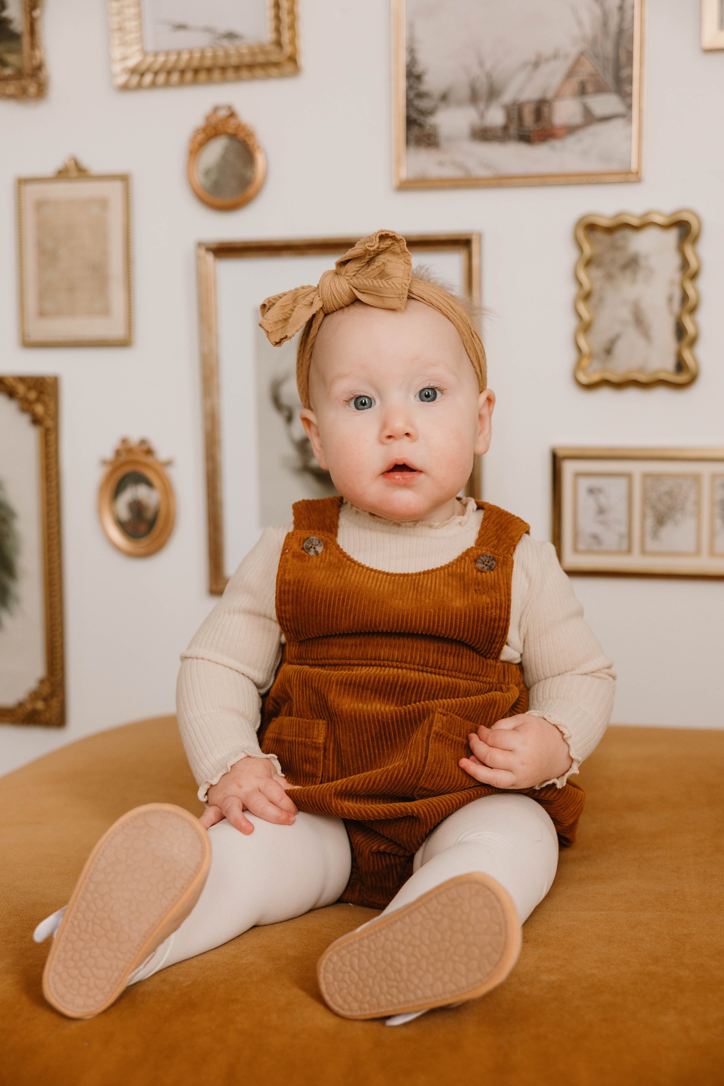A baby girl with a head wrap, dressed in a tan long-sleeve shirt and rust-colored corduroy dress, sitting on a mustard yellow surface with a gallery wall of various framed pictures behind her.