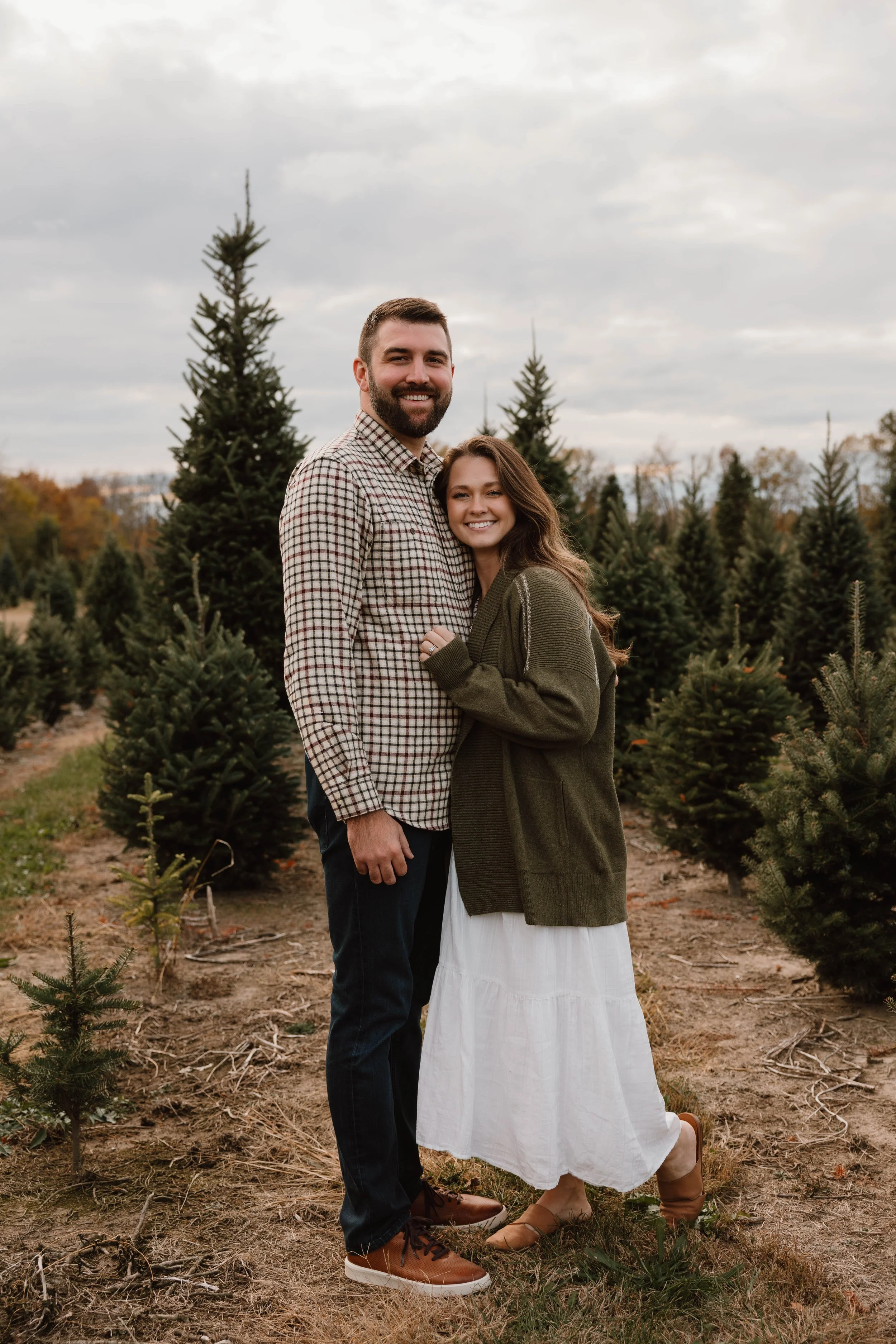 A couple standing together in a Christmas tree farm, smiling at the camera, surrounded by small evergreen trees and overcast sky.