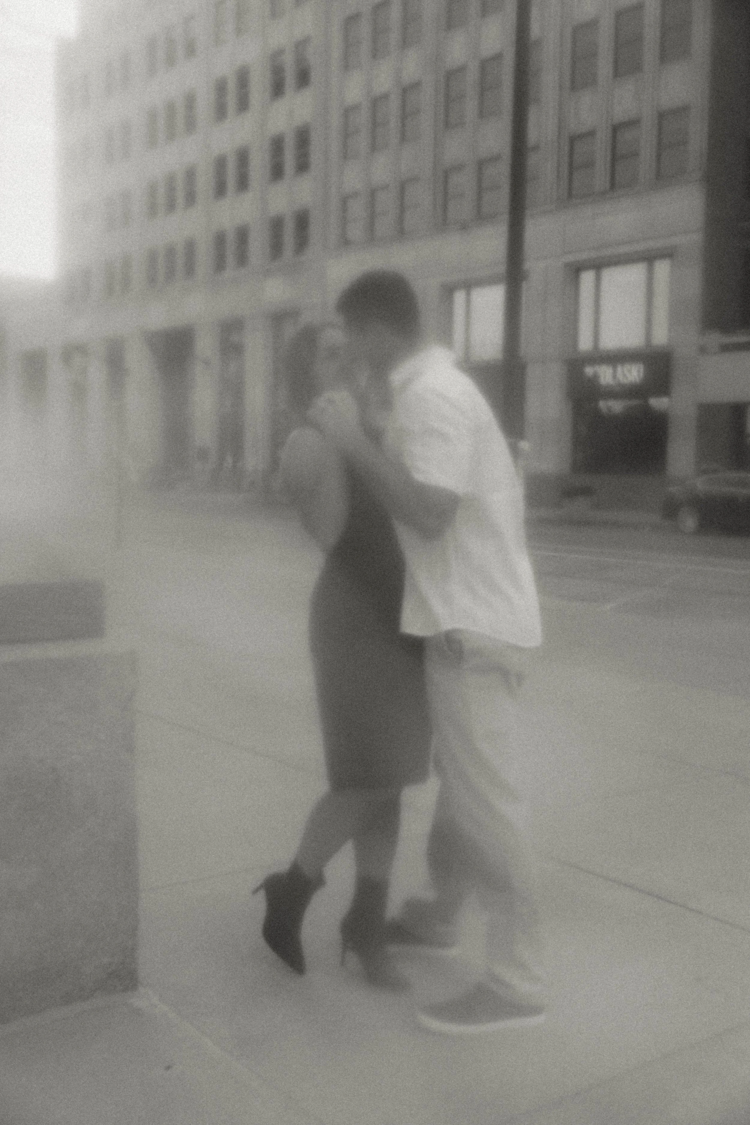 A black and white photo of a couple kissing on a city sidewalk, with tall buildings and parked cars in the background.