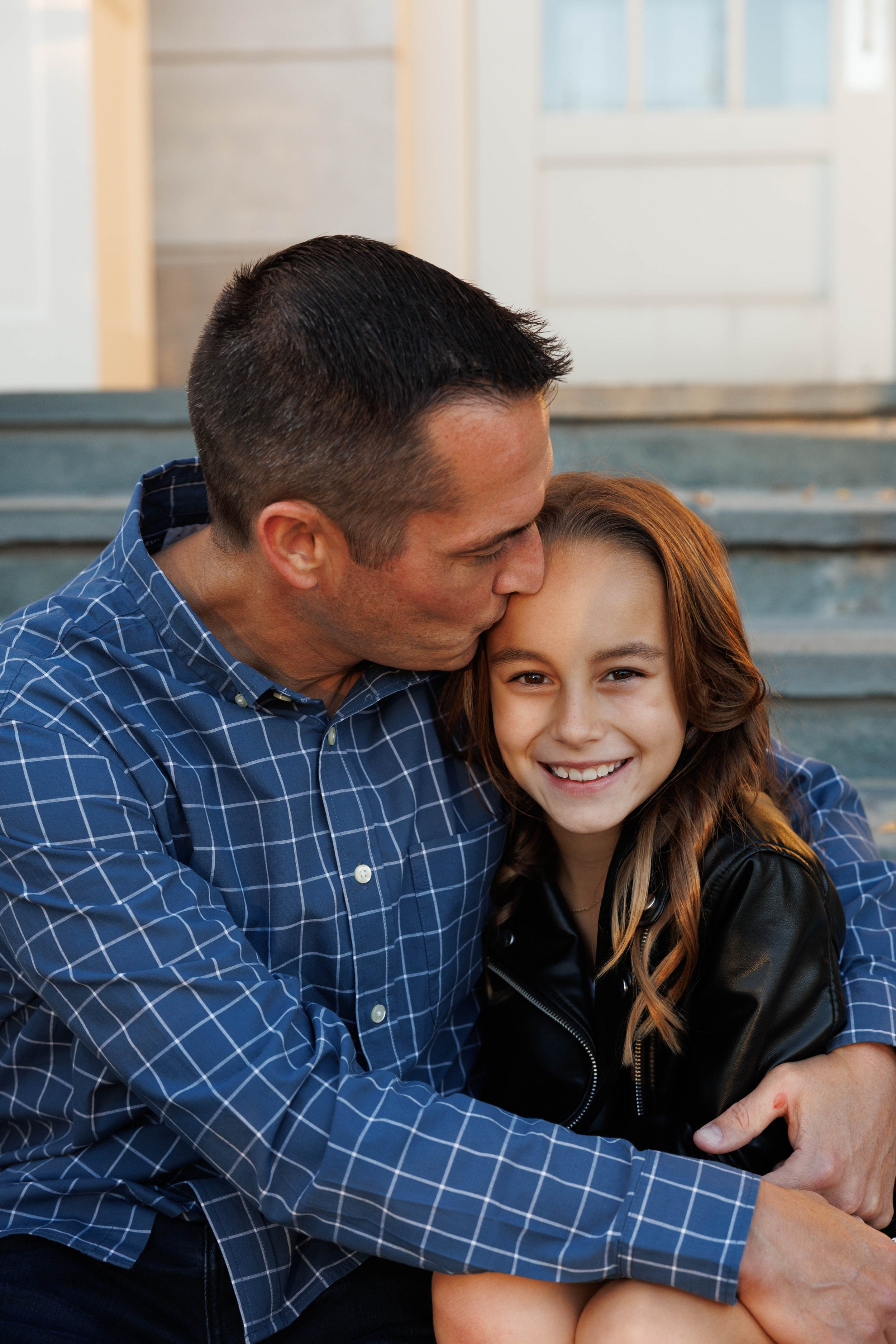 A man with short dark hair wearing a blue checkered shirt hugging and kissing a smiling girl with long brown hair in a black leather jacket sitting on steps outside a house.