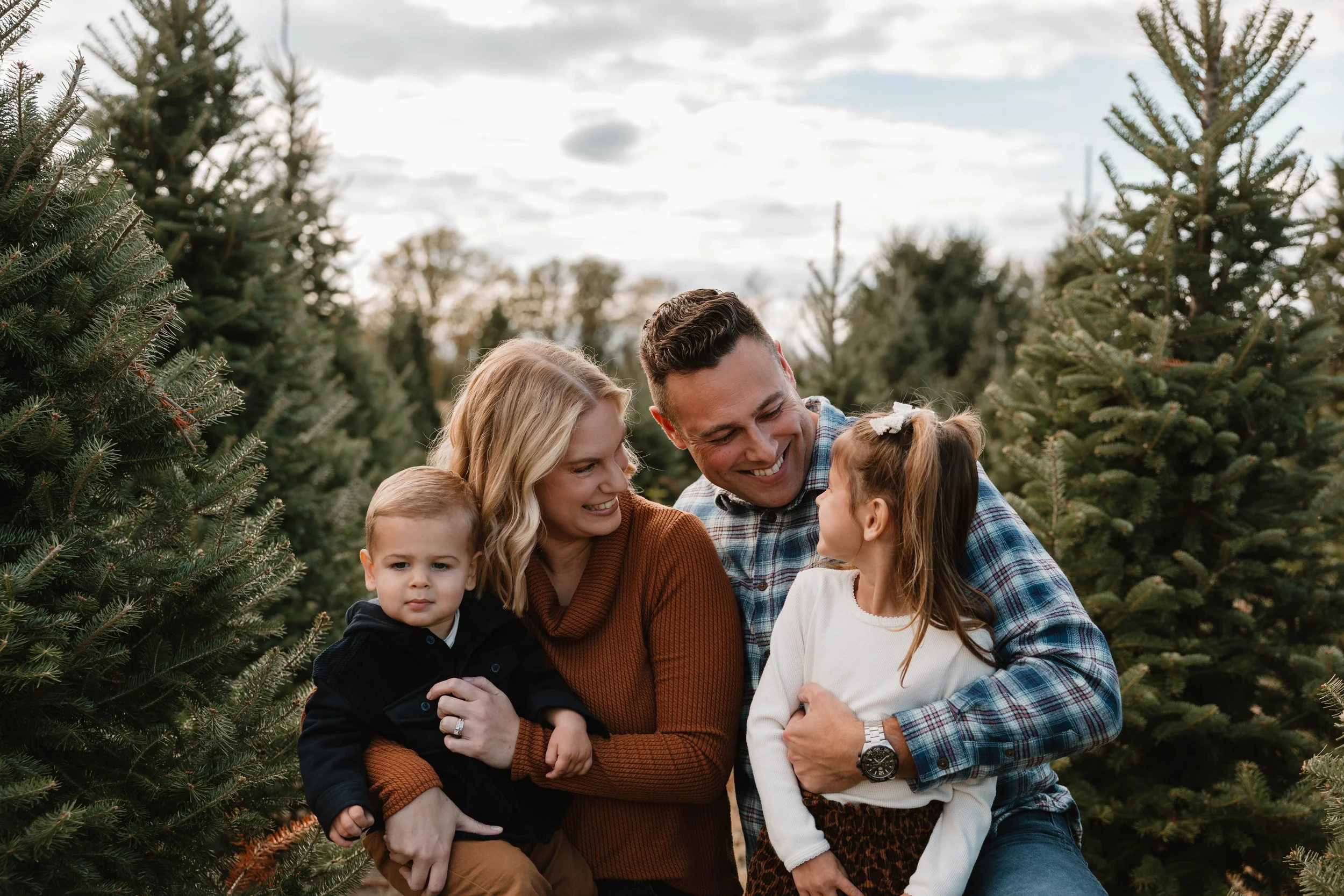 A family of four, including a man, woman, and two children, smiling and enjoying time together among Christmas trees outdoors during the daytime.