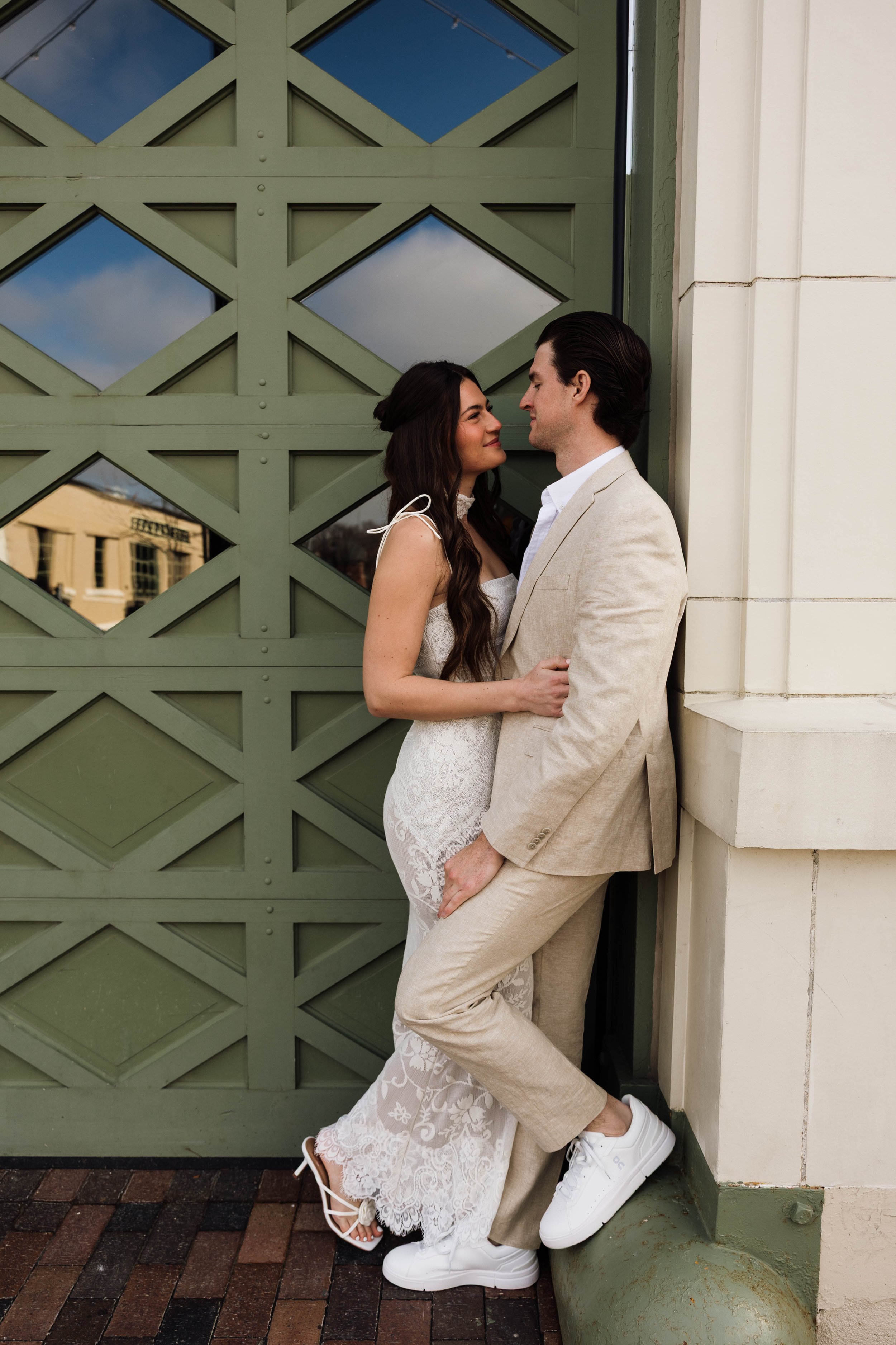 A couple dressed in wedding attire, standing close and facing each other, outdoors in front of a green geometric-patterned door. The woman wears a white lace wedding dress and the man is in a beige suit with white sneakers.